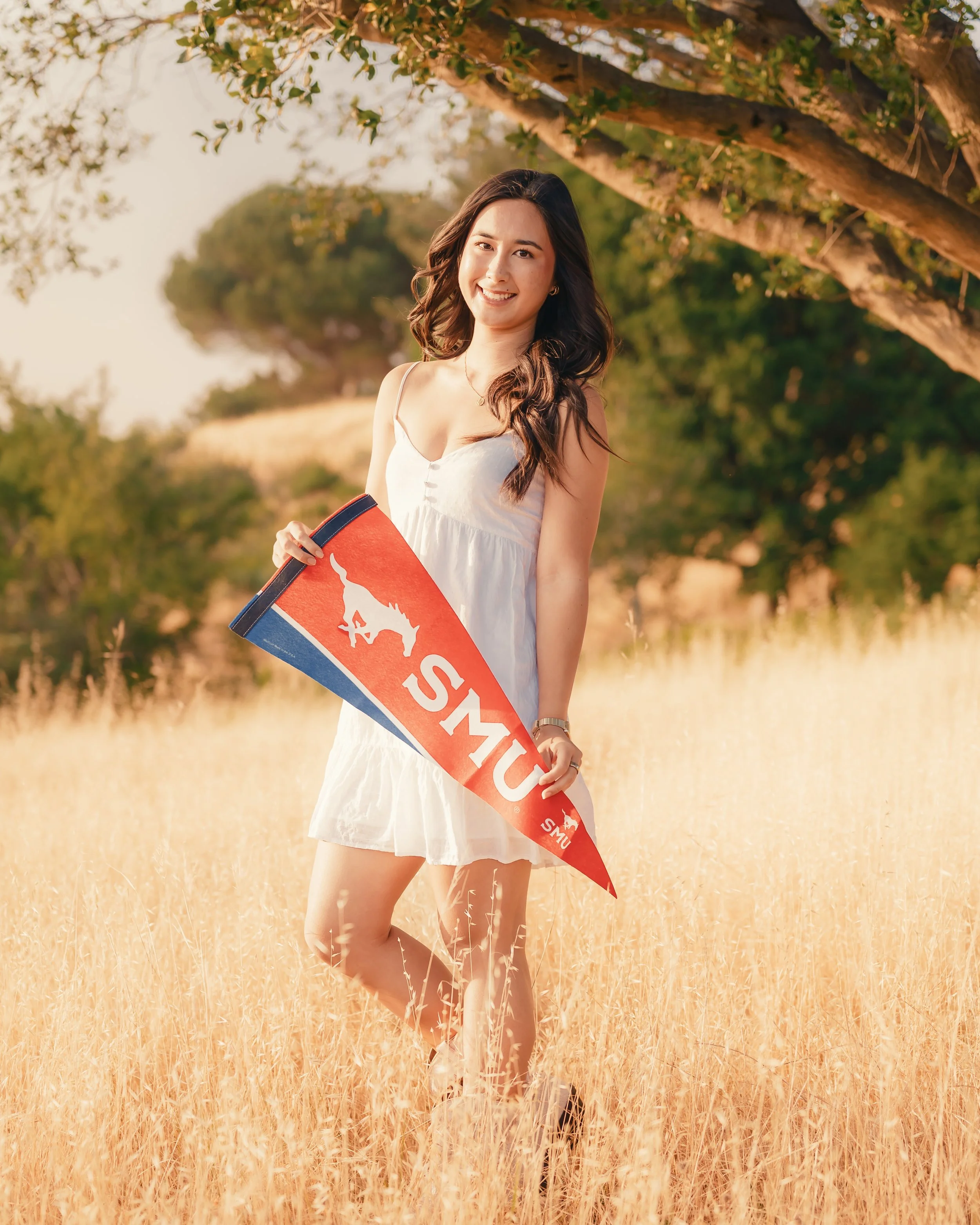 A young woman with long dark hair, wearing a white dress, smiling and holding a red and blue foam finger with a logo, standing in a field of tall dry grass with green trees and a clear sky in the background.