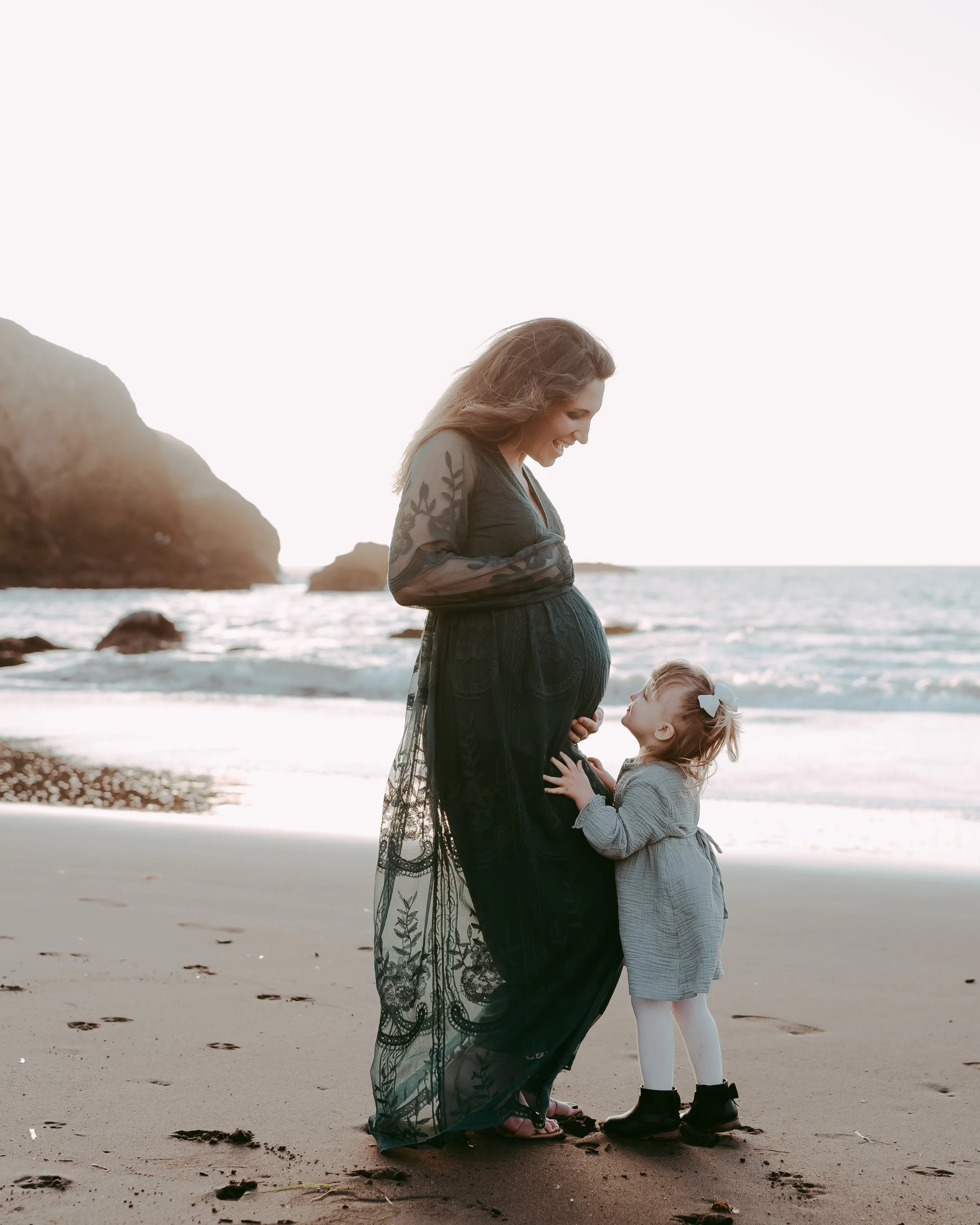 A pregnant woman standing on a beach with a little girl looking up at her, gently touching her baby bump as they smile at each other during sunset.