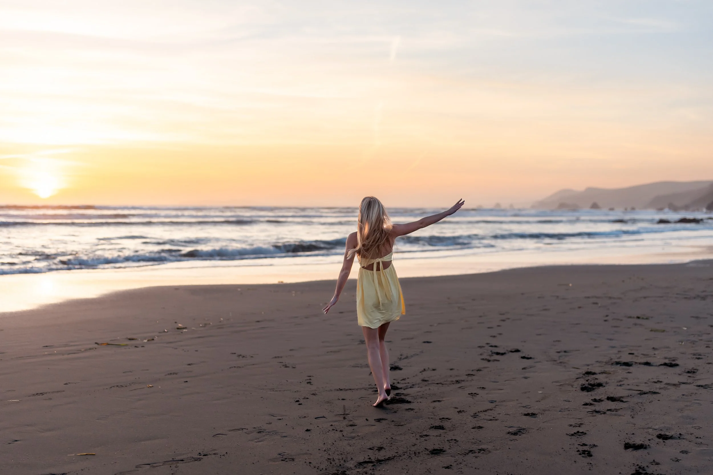 A woman with long blonde hair in a yellow dress walking barefoot on the beach during sunset.