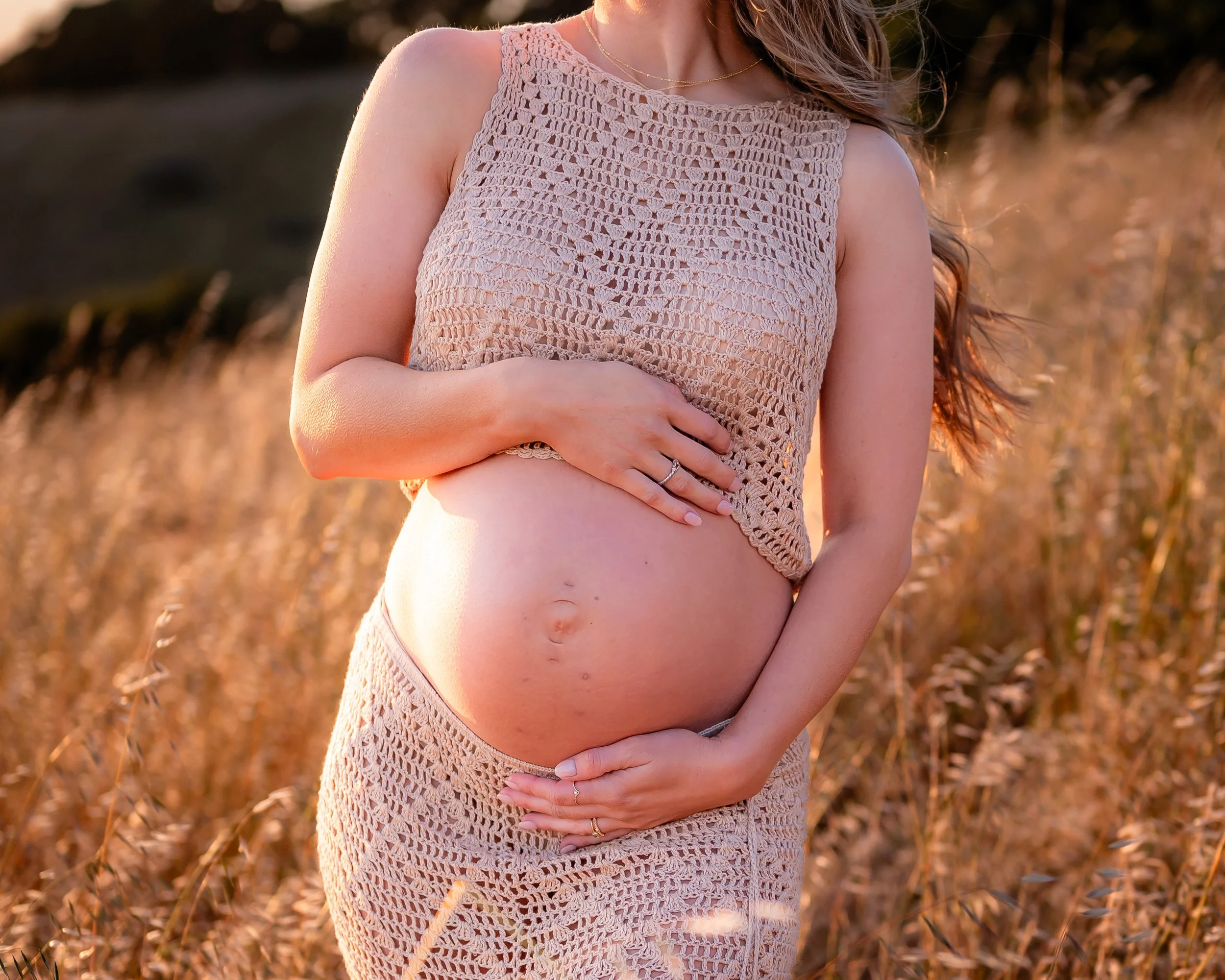 A pregnant woman wearing a crochet top and skirt standing in a field of tall grass at sunset, gently holding her belly.