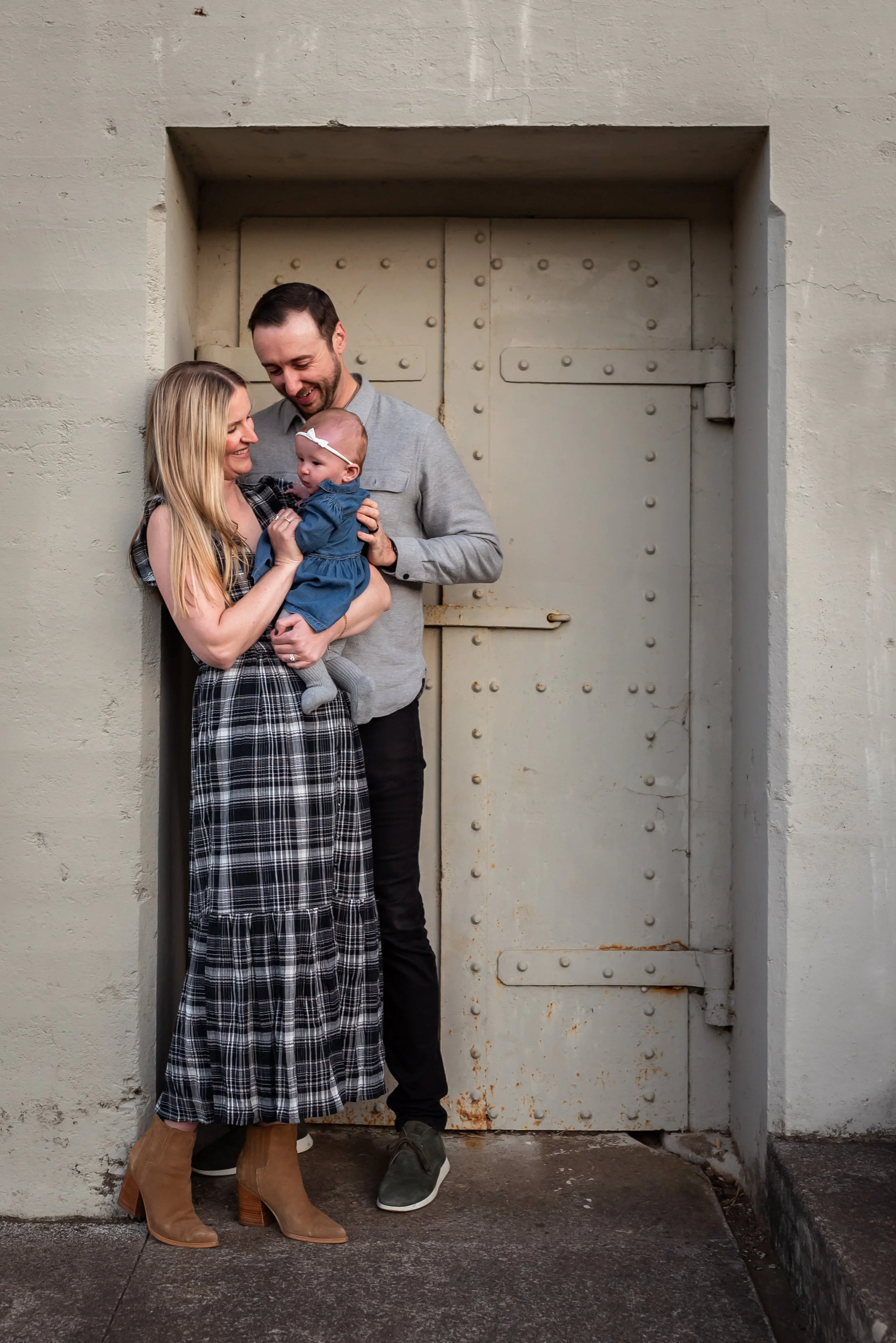A smiling woman, man, and baby girl standing in front of a large metal door. The woman is holding the baby, and the man is close to them, all looking at each other happily.
