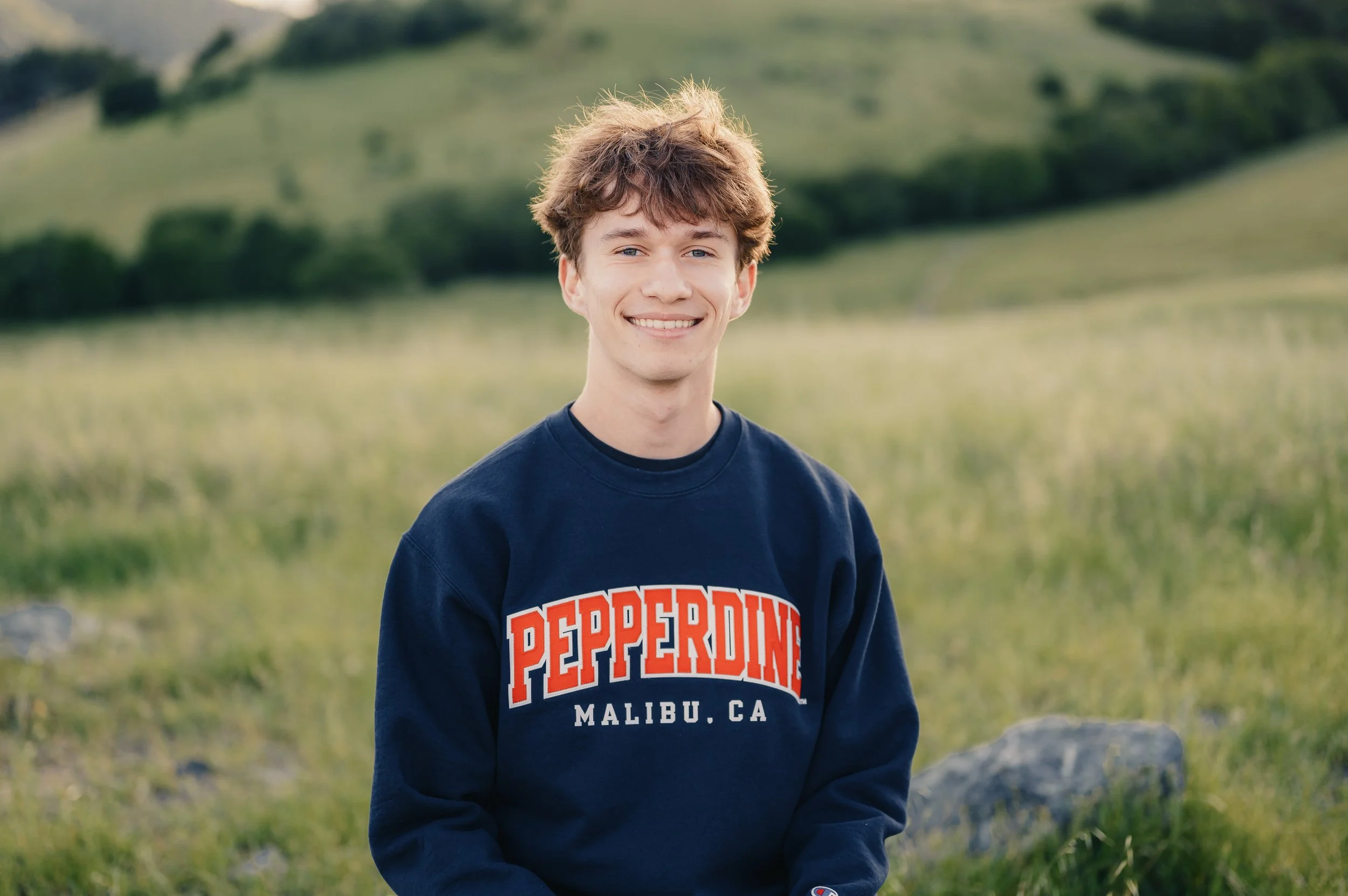 Young man with light brown hair smiling outdoors, wearing a navy blue sweatshirt with red and white text, in a green landscape with trees and rocks.