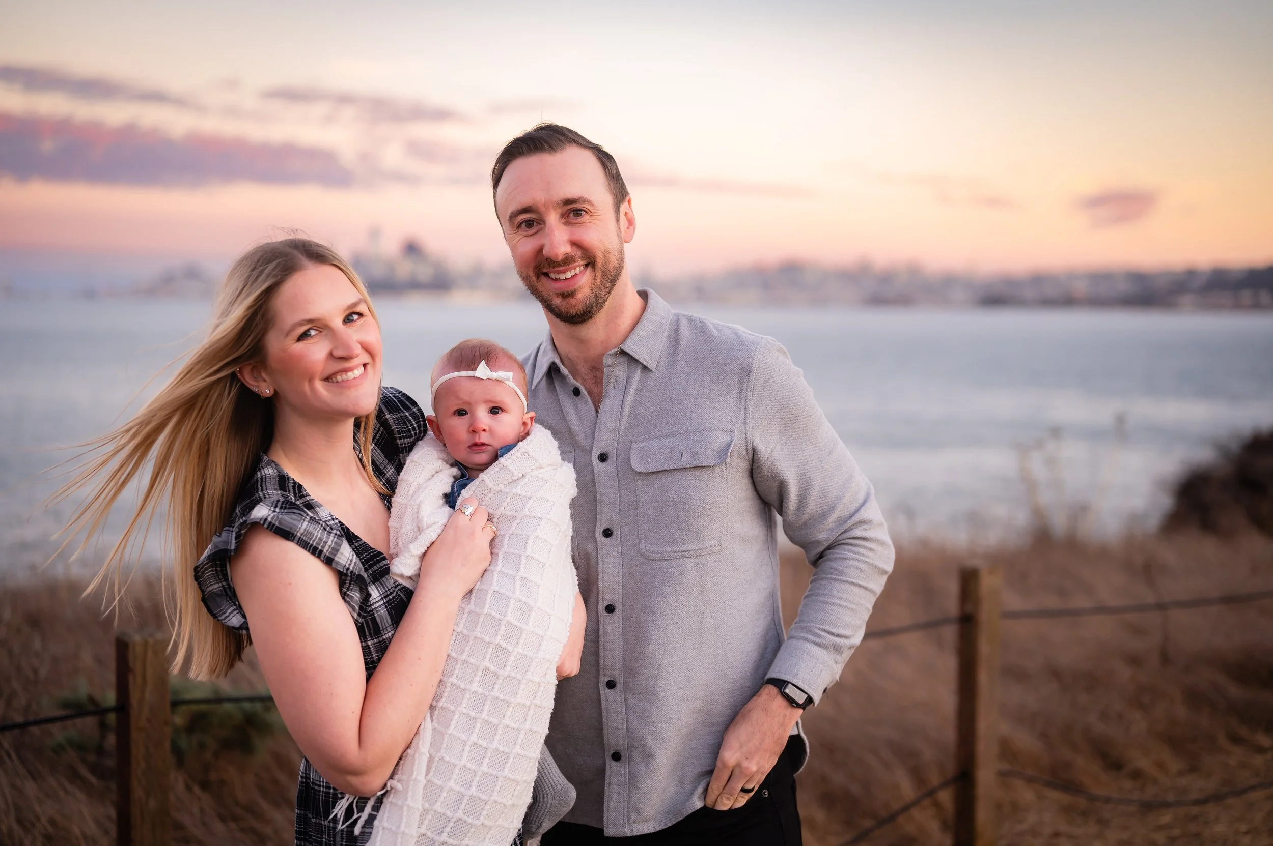 A smiling family of three standing outdoors near the coast during sunset, with water and city skyline in the background. The mother is holding a baby girl wrapped in a blanket, and the father is standing beside them.