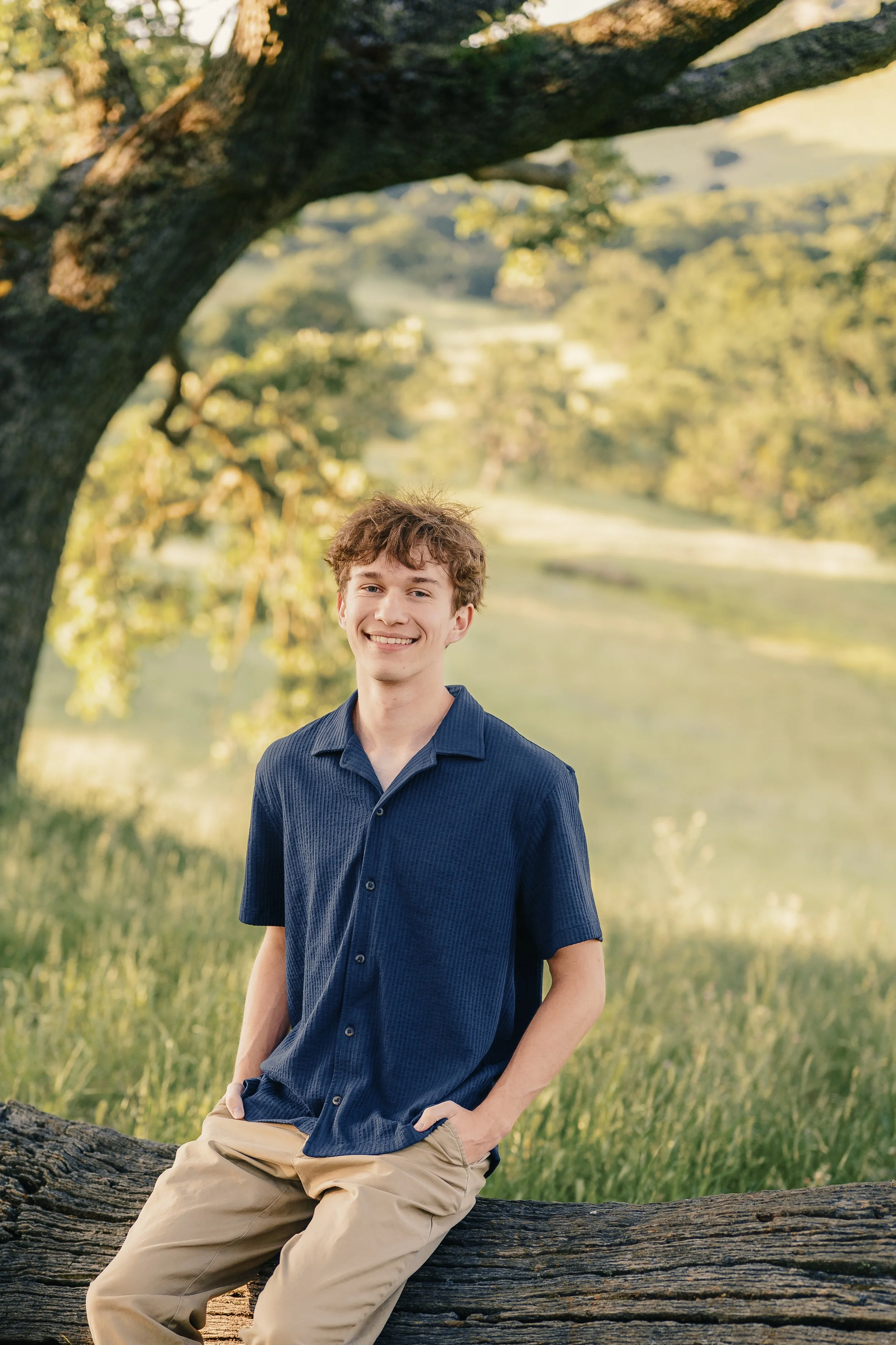 A young man with brown curly hair and a blue button-up shirt, smiling and standing outdoors near a large tree and a body of water, during daytime.