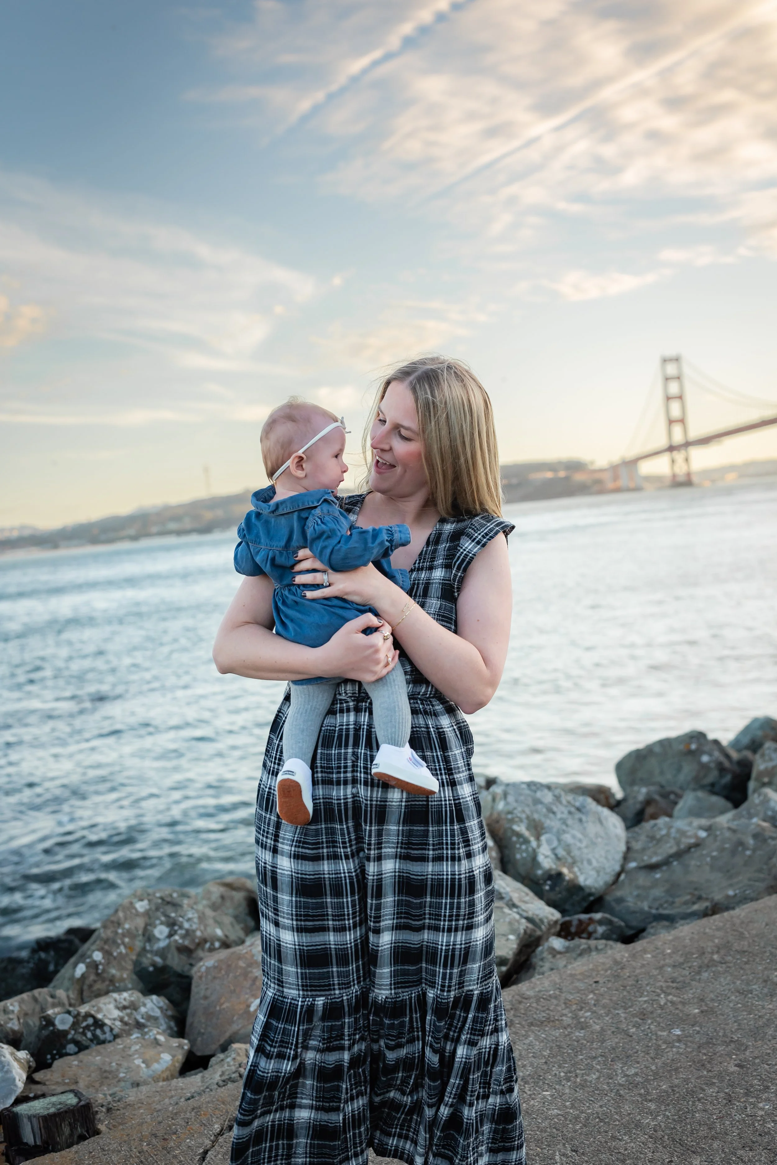 A woman with blonde hair holding a baby girl by the water with the Golden Gate Bridge in the background during sunset.