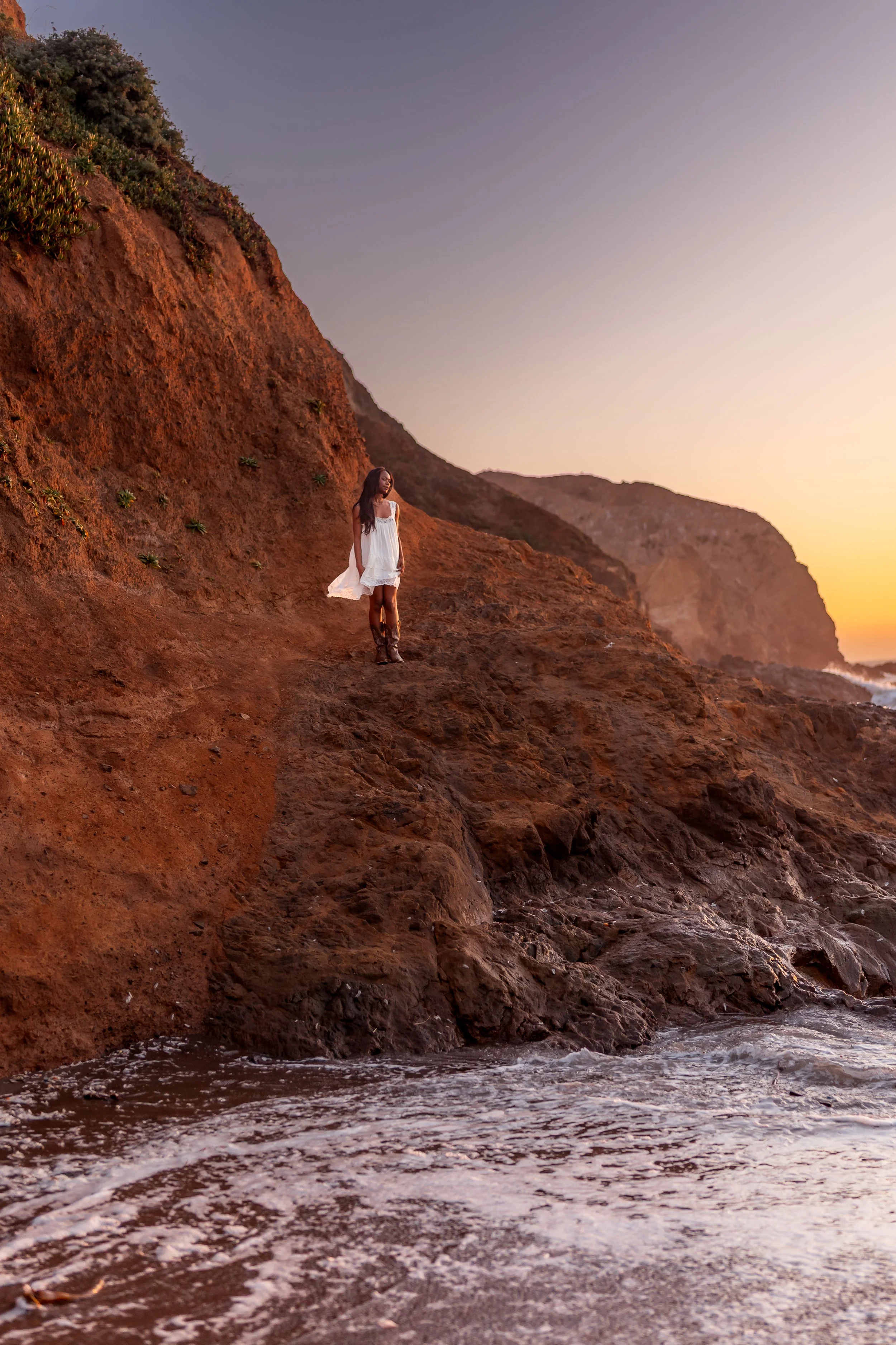 A woman in a white dress and cowboy boots standing on rocky cliffside at sunset, overlooking the ocean with waves below.