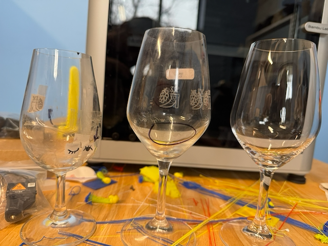 Three empty wine glasses on a wooden table, with a black device and colorful craft supplies, like yellow and blue straws, scattered around. In the background, there is a computer monitor.
