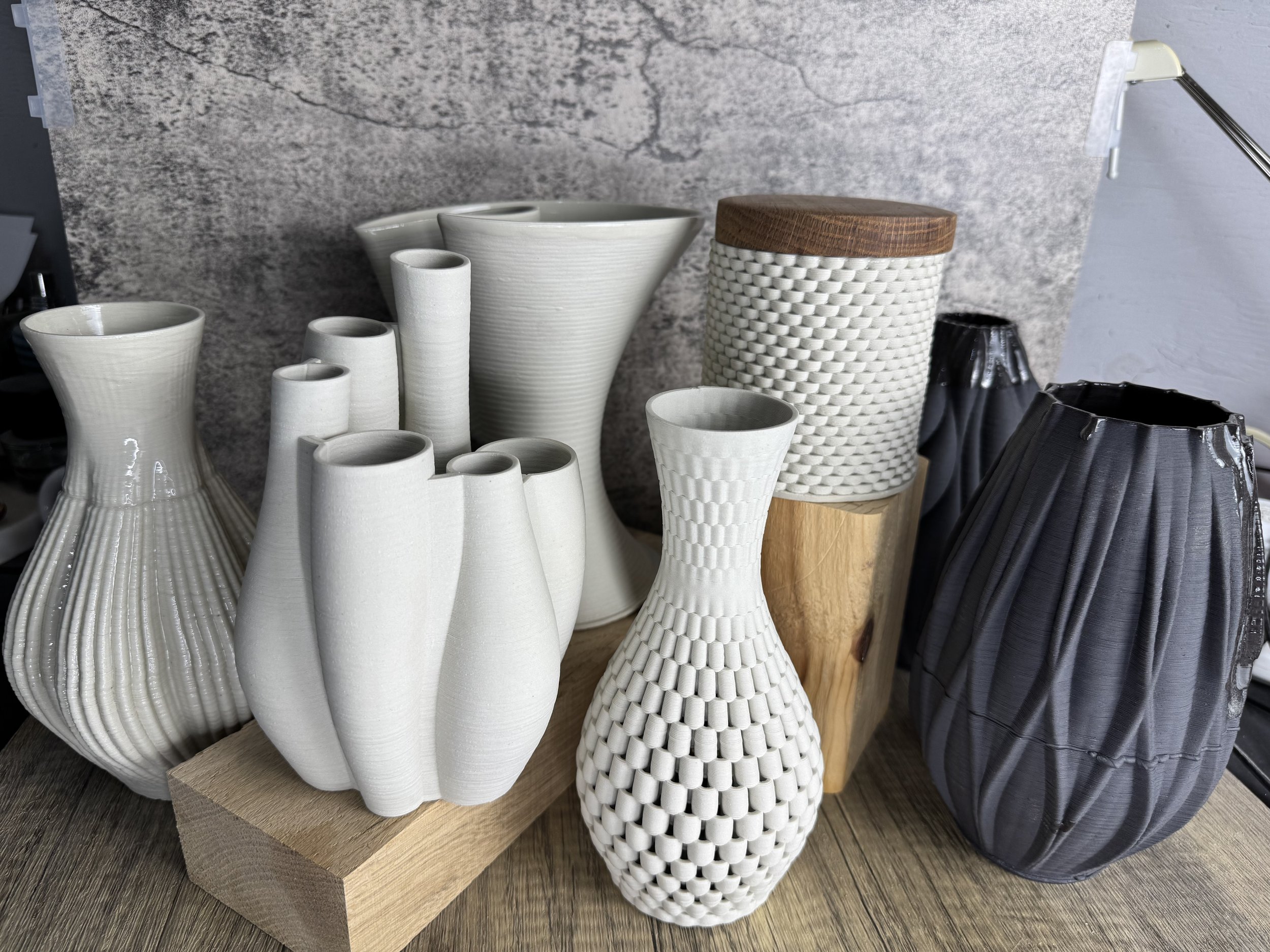 A display of various vases and containers made of ceramic and wood, arranged on a wooden surface against a textured gray wall.