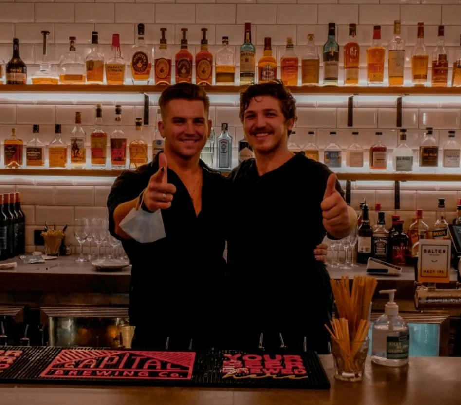 Two smiling men behind a bar counter in a bar, giving thumbs up, with bottles of alcohol on shelves behind them.
