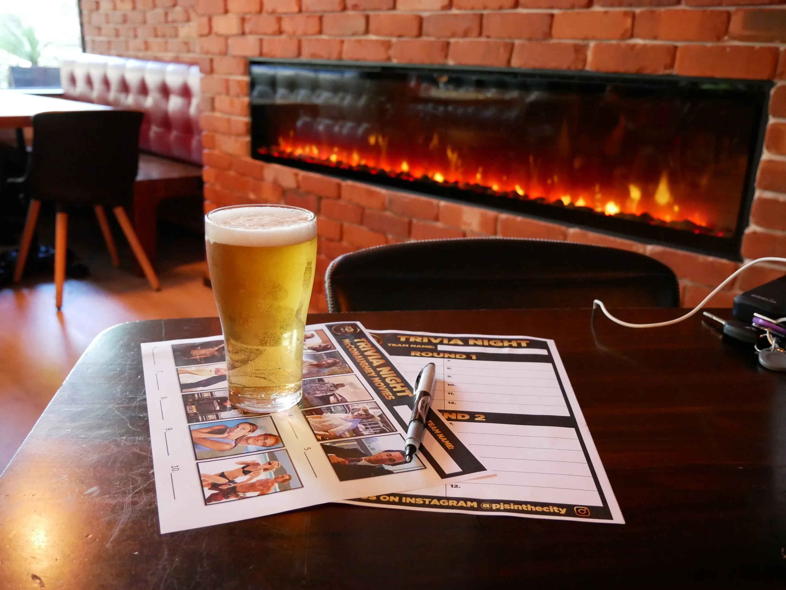 A table with a glass of beer, a pen, and a trivia night sheet in a cozy restaurant with a red brick wall and a fireplace in the background.