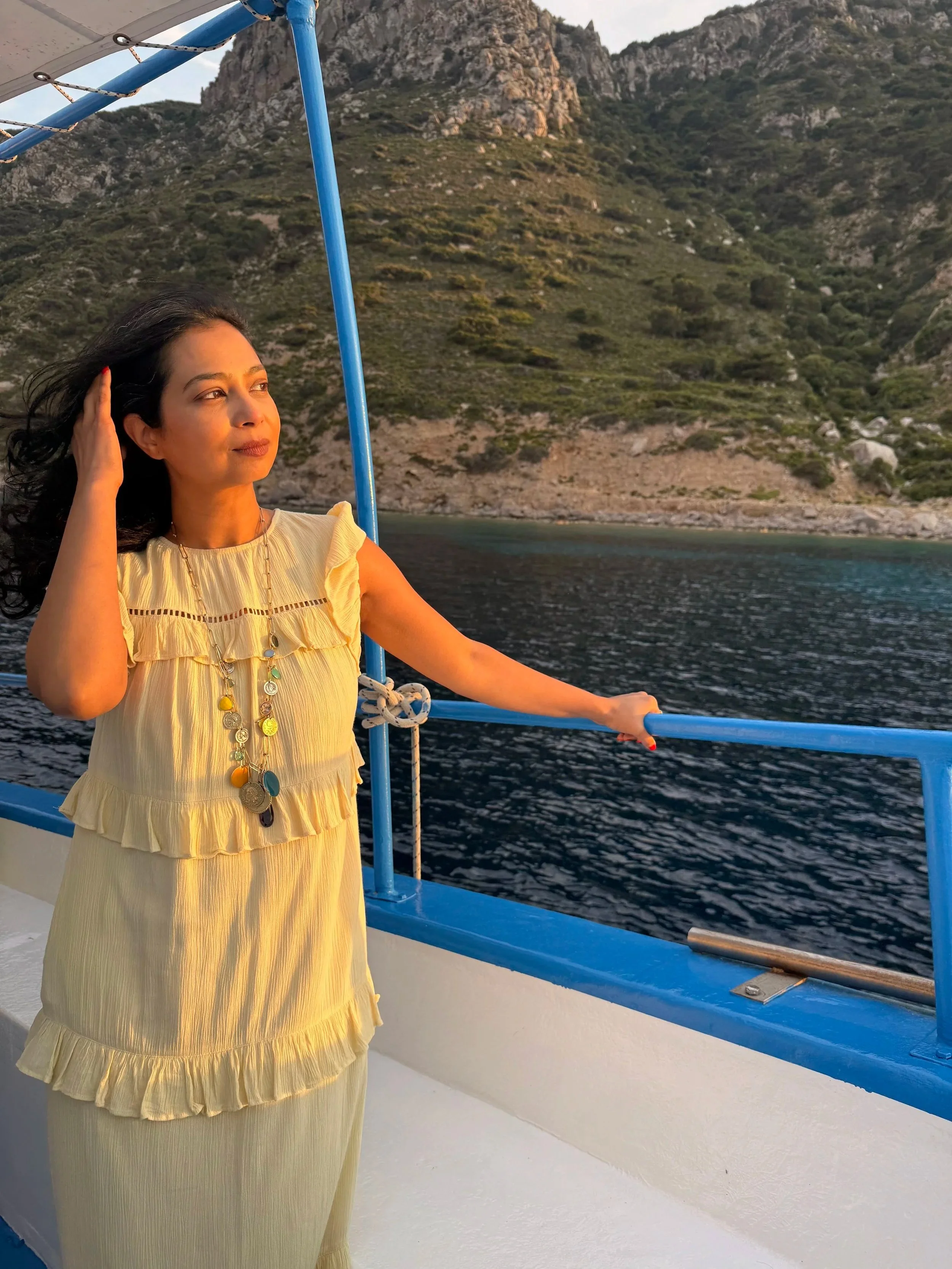 Woman in a yellow dress standing on a boat, gazing into the distance with her hand touching her hair, overlooking a coastal landscape with water and green hills.
