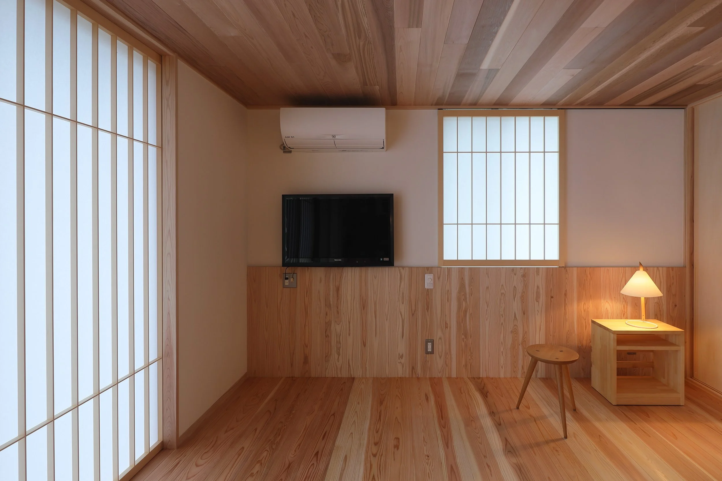 木の腰壁と白い壁にはテレビとエアコンが設置されています。内障子に守られた空間にはスツールと照明器具が置かれている。