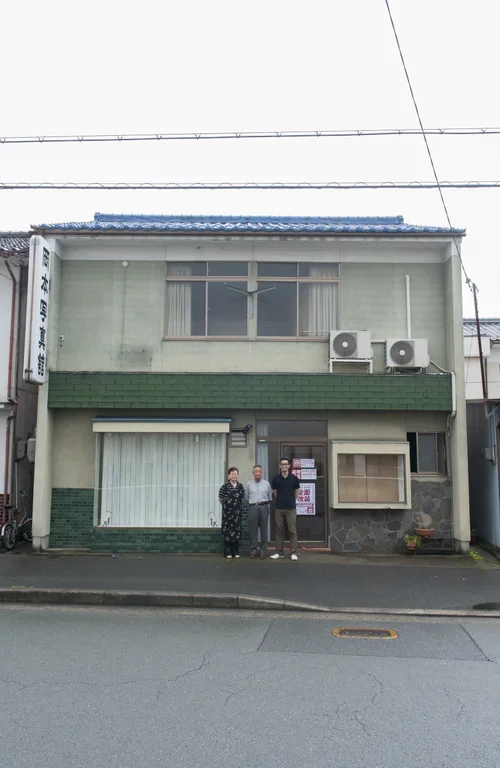 緑の壁面に青い屋根の建物の前に３人の人が建っている