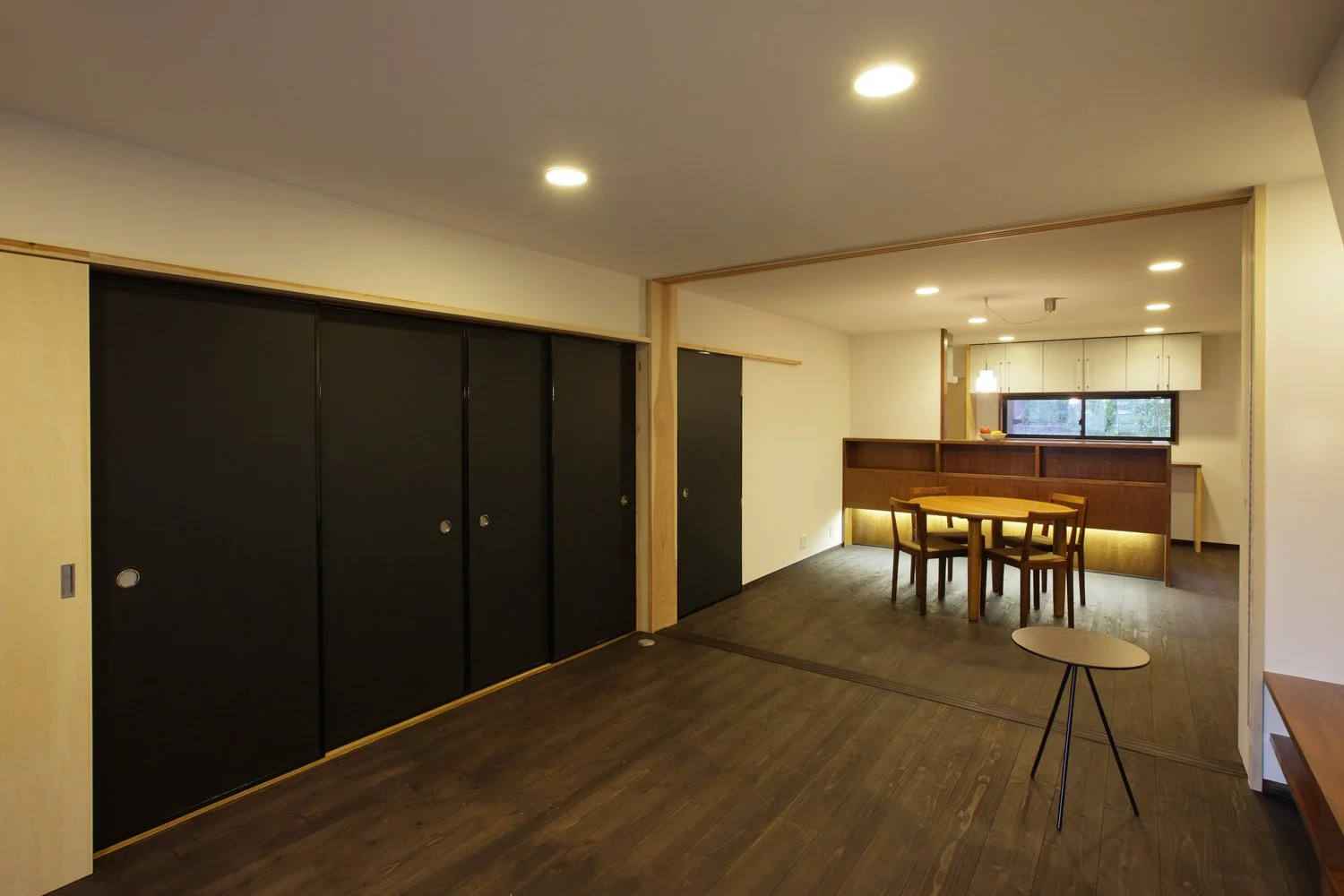 白い壁と天井、黒い床に黒い襖で仕切られた空間。部屋の片隅に置かれた丸いテーブルと椅子の奥にカウンターが見えている。