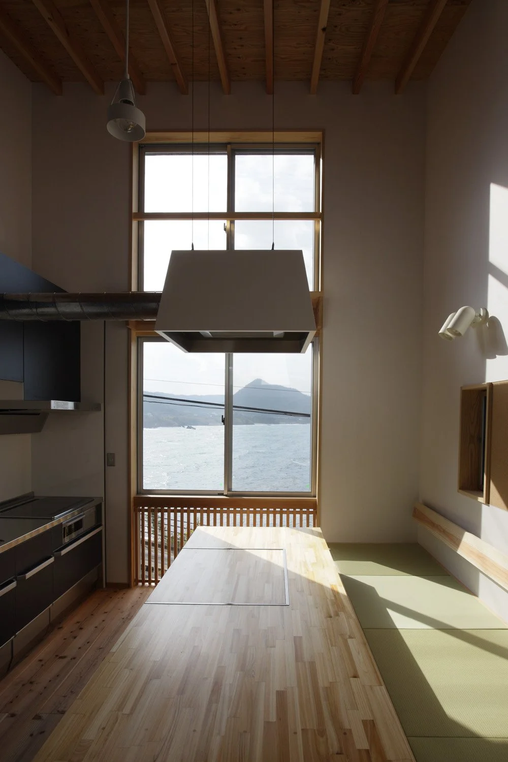 縦長の窓の外には海が見える部屋。テーブルの上には台形の換気フードが吊り下げられている。部屋の右端は畳敷きの座敷があり、左端にはキッチンがある。