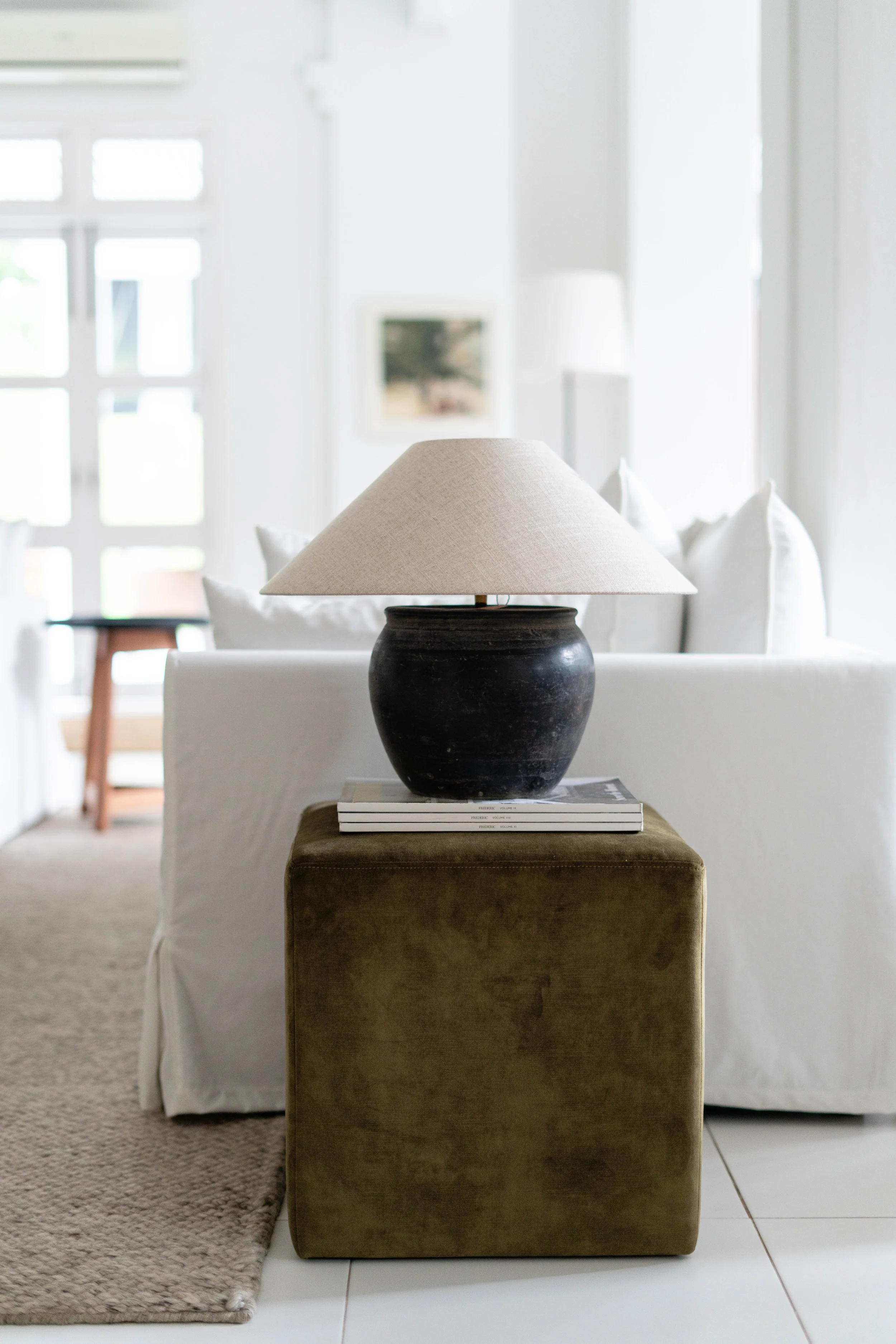 A living room with a black vase holding a beige lampshade, placed on a small green ottoman with a stack of books underneath, white curtains and chairs in the background.