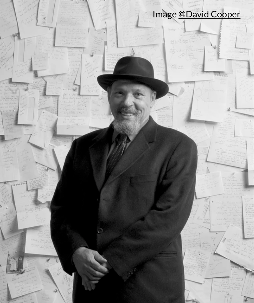 Man wearing a dark suit and hat, standing in front of a wall covered in different sized paper and notes.  Black and white image by David Cooper.
