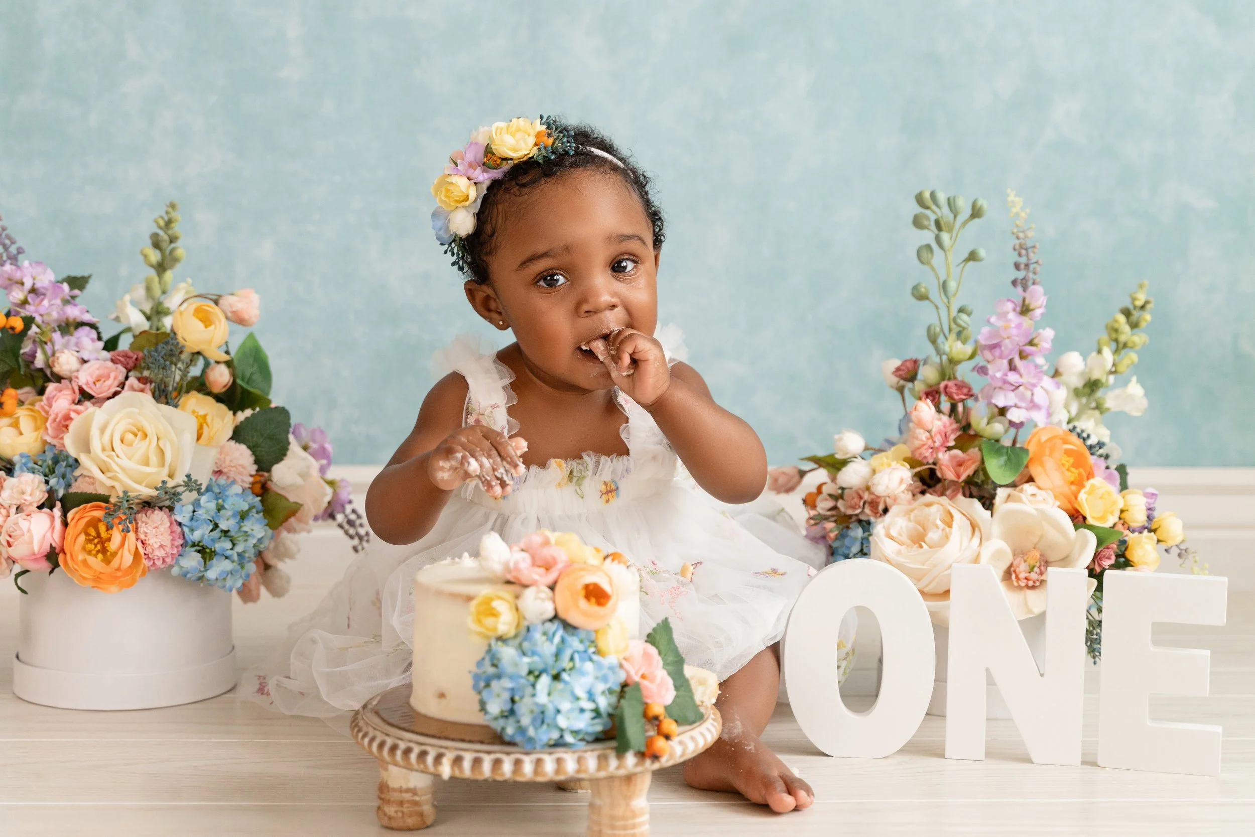 Baby girl celebrating her first birthday with a cake smash, surrounded by soft floral decorations made by her mom
