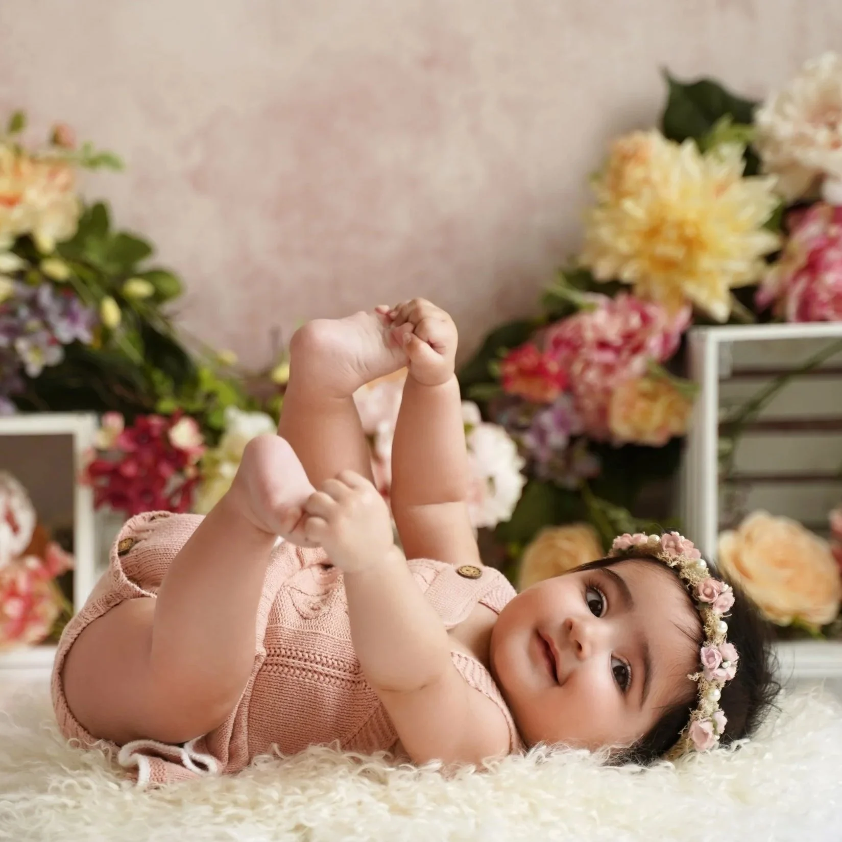 Milestone photos with 6 month old girl, with a big smile, lying on her side on a soft, cream-colored rug, looking directly at the camera and grabbing her toes. She wears a crown of pink and cream flowers and a light pink dress.