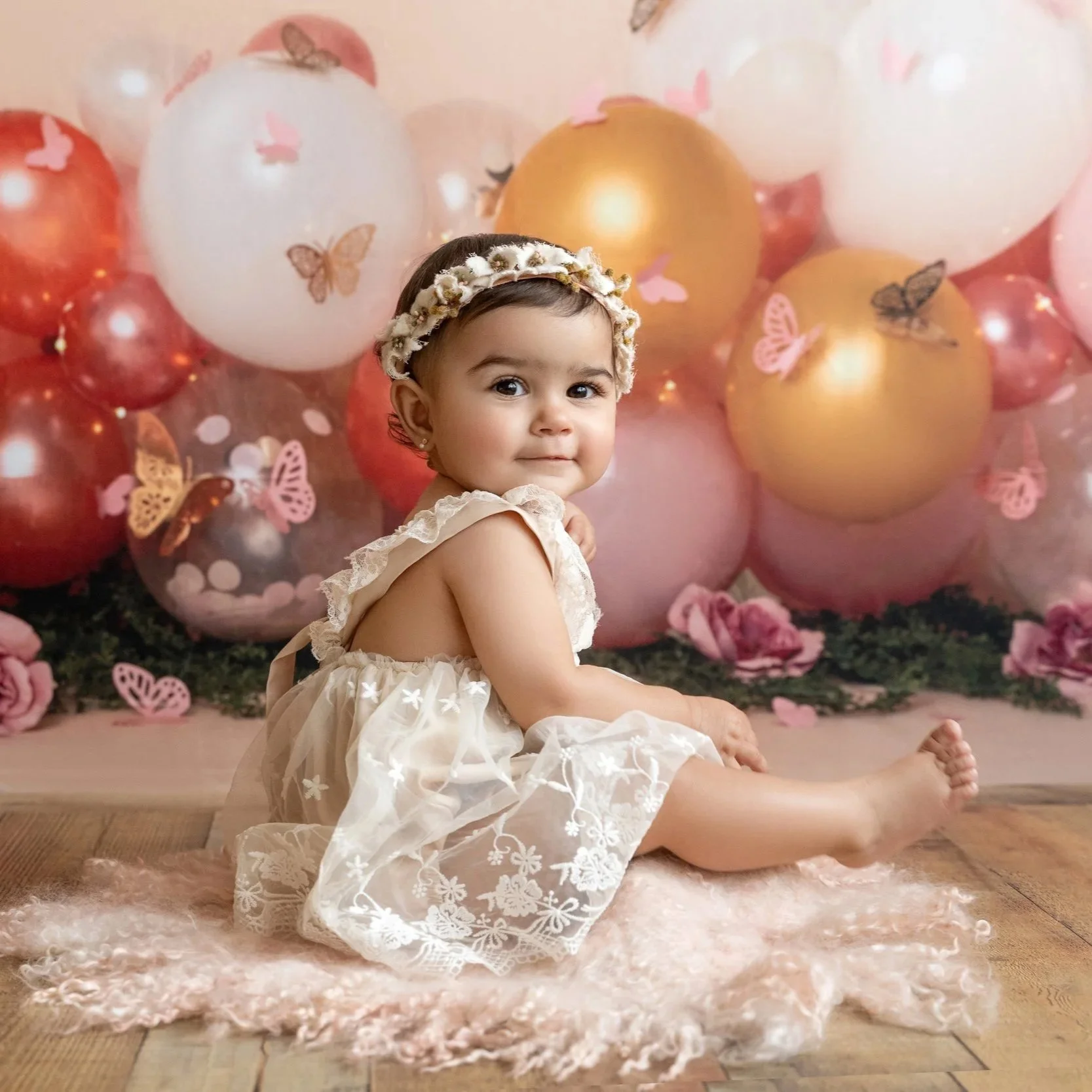 Birthday photos with one year old girl who is sitting on a fluffy pink rug with young girl, surrounded by pink and gold balloons decorated with butterflies. Getting ready for her cake smash.