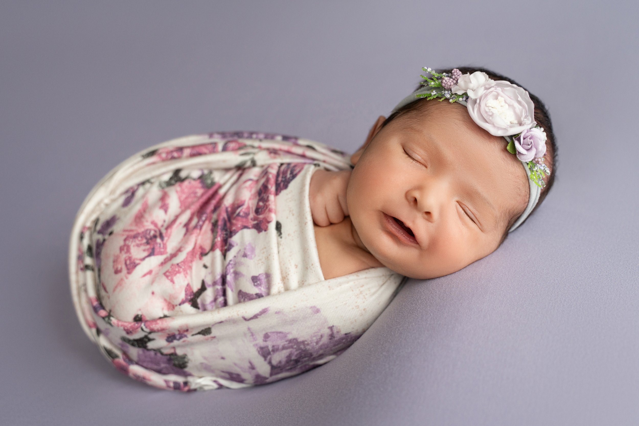 Newborn smiling while sleeping and swaddled in pink and purple flower wrap
