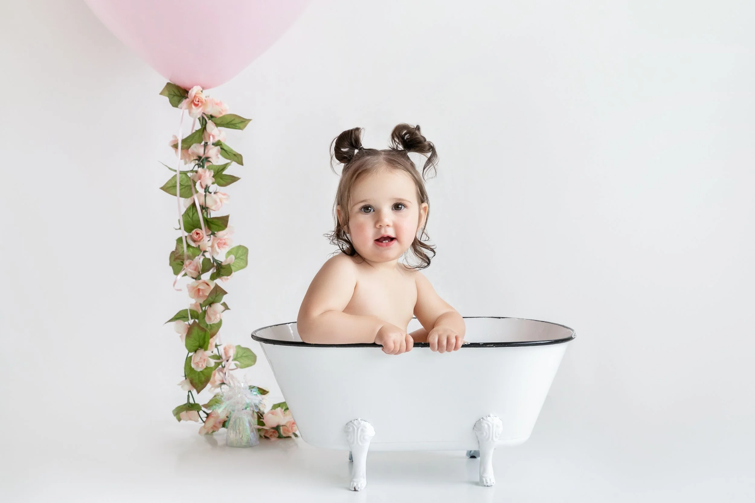 One-year-old girl sitting in a vintage clawfoot bathtub after her cake smash milestone photography session with pink balloon decor.