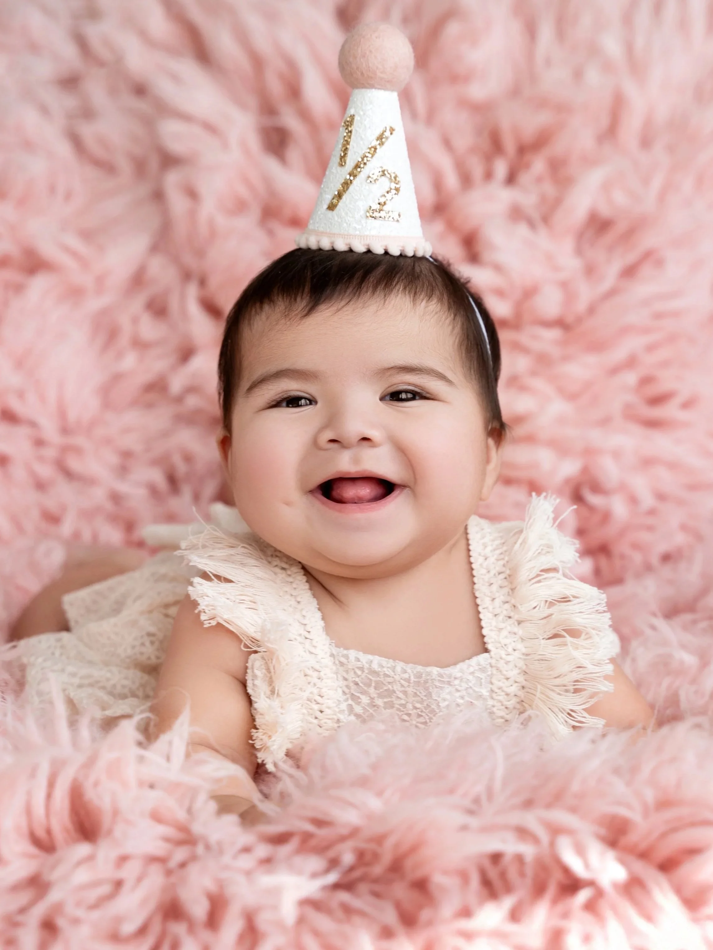 A smiling baby girl at her 6 month photo session wearing a white dress lying on a pink fluffy blanket, with a birthday hat on her head that says '1/2' in gold glitter, with a large pink pom-pom on top.