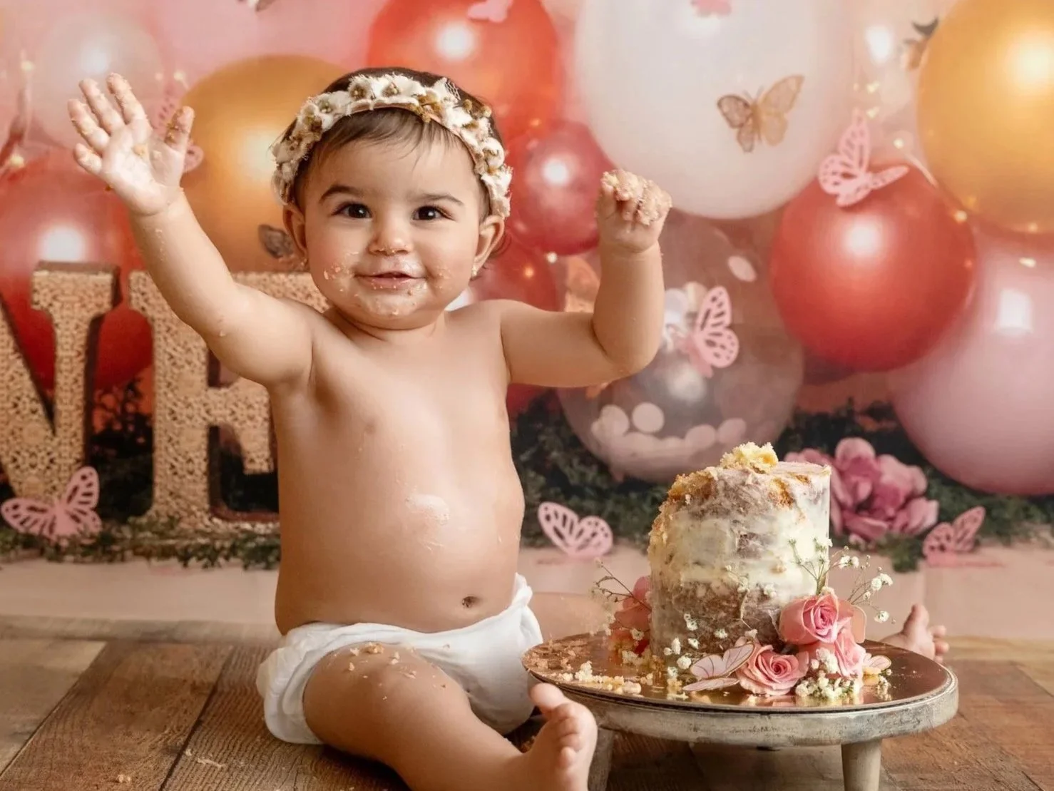 A one-year old baby girl at her Cypress cake smash photo session with cake and frosting on face and body. The baby has a floral headband and is raising arms happily. The background features pink, peach, and white balloons, and butterflies.