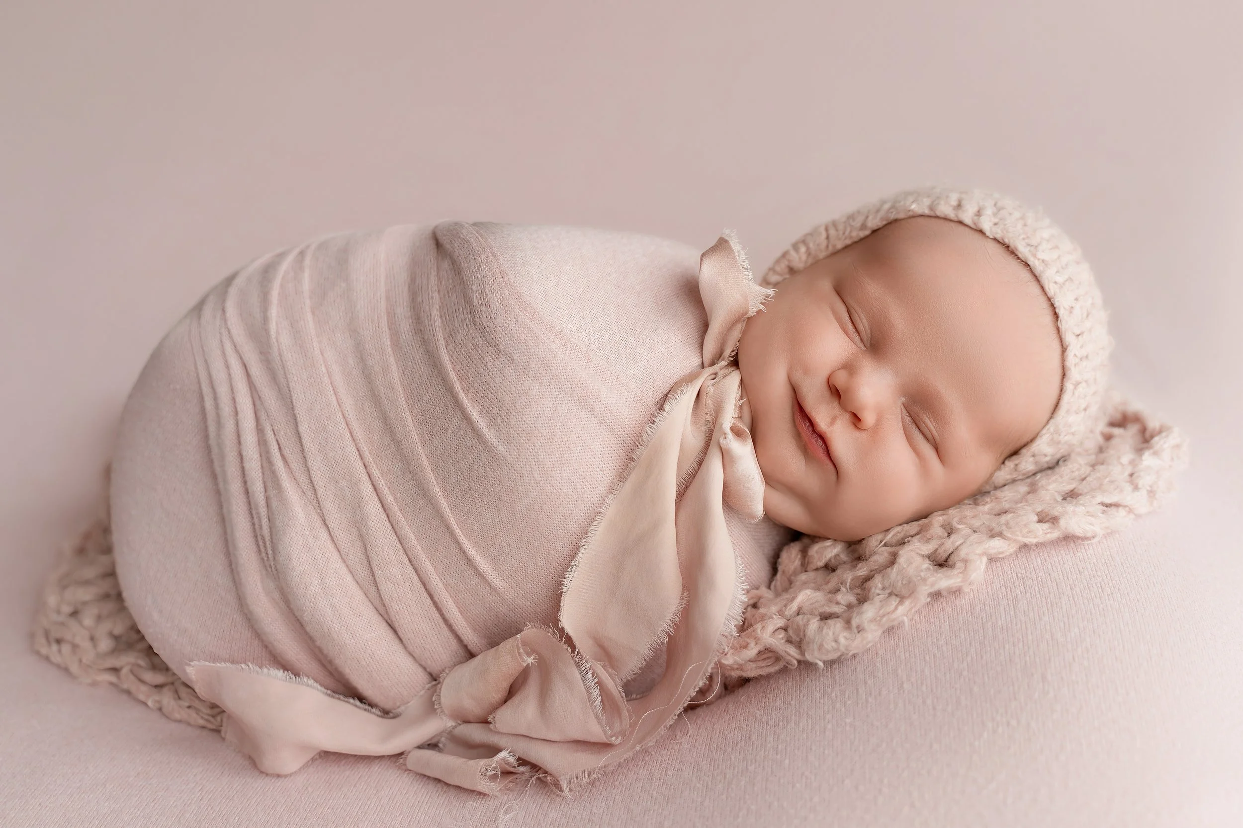 Newborn baby girl peacefully wrapped in soft blush pink during a posed newborn photo session.