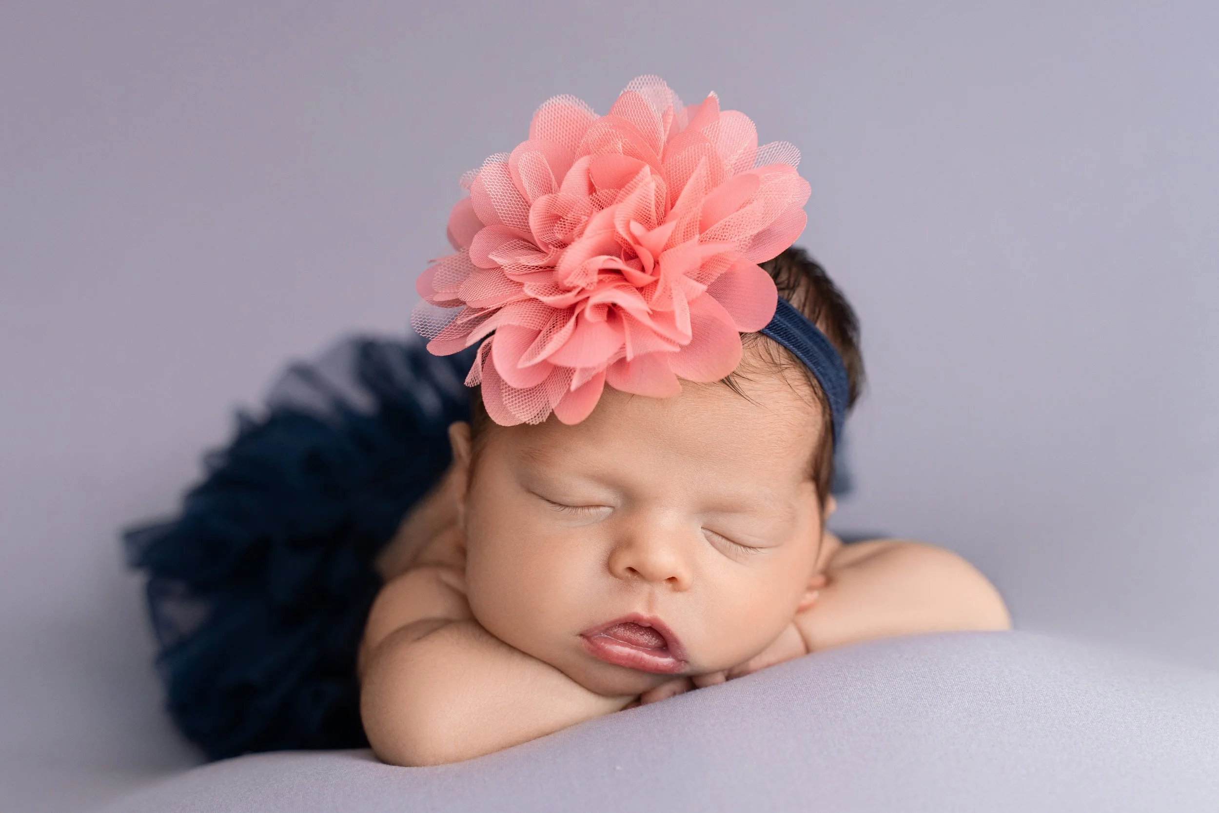 Close-up portrait of newborn baby Jocelyn sleeping with her arms folded beneath her chin and a large pink flower headband.