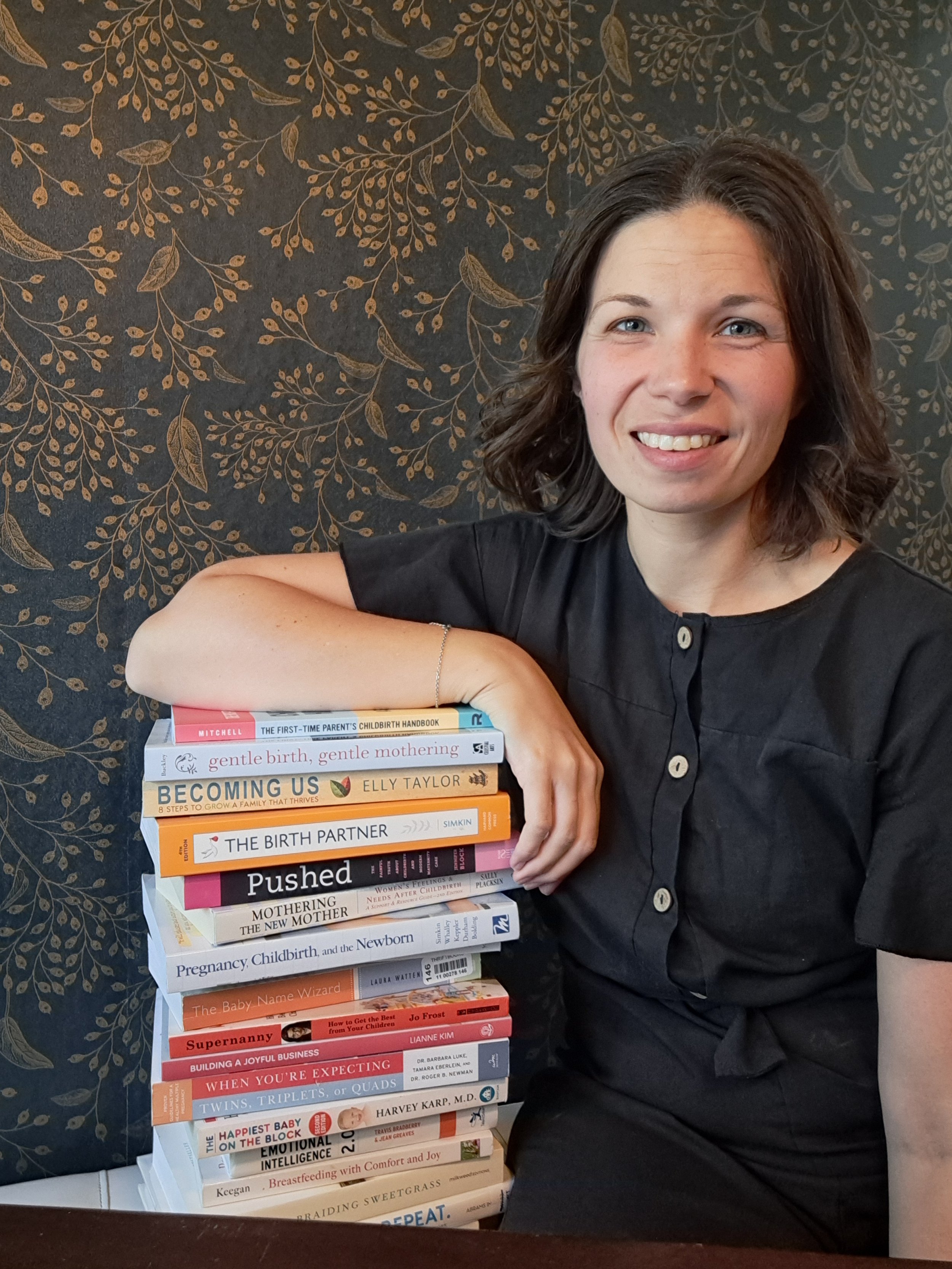 A woman with shoulder-length brown hair and a black shirt sitting next to a stack of books about pregnancy, childbirth, and parenting, smiling at the camera against a dark floral-patterned background.