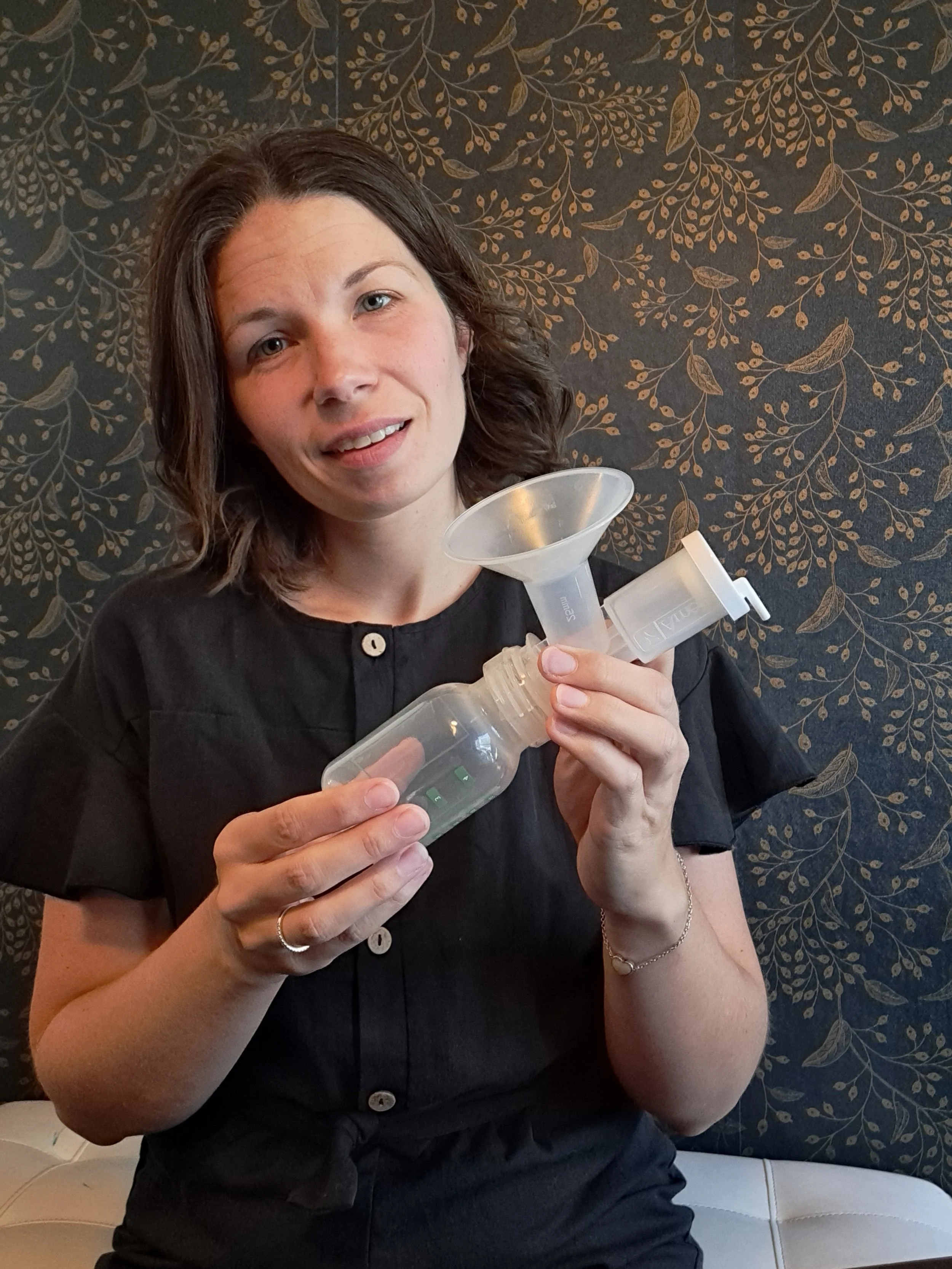 A woman with shoulder-length dark hair holding a breast pump, sitting against a dark, floral-patterned wall.