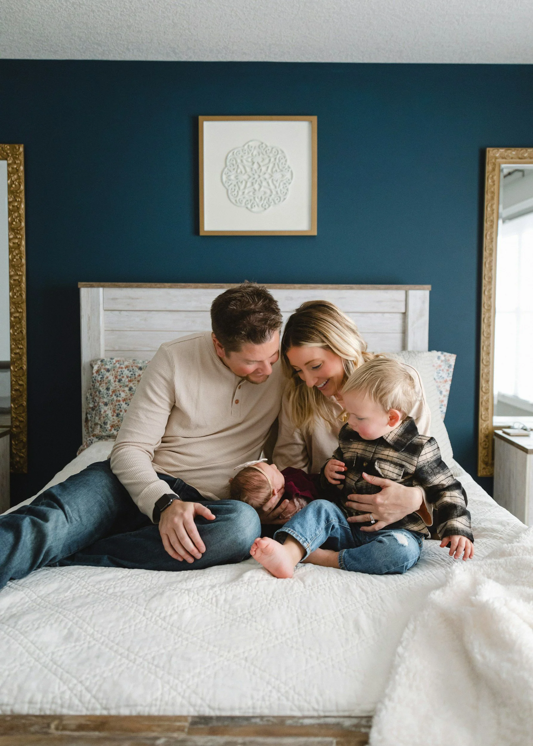 Family of four sitting on a bed, smiling and holding their newborn baby, in a cozy bedroom with blue walls and framed artwork.
