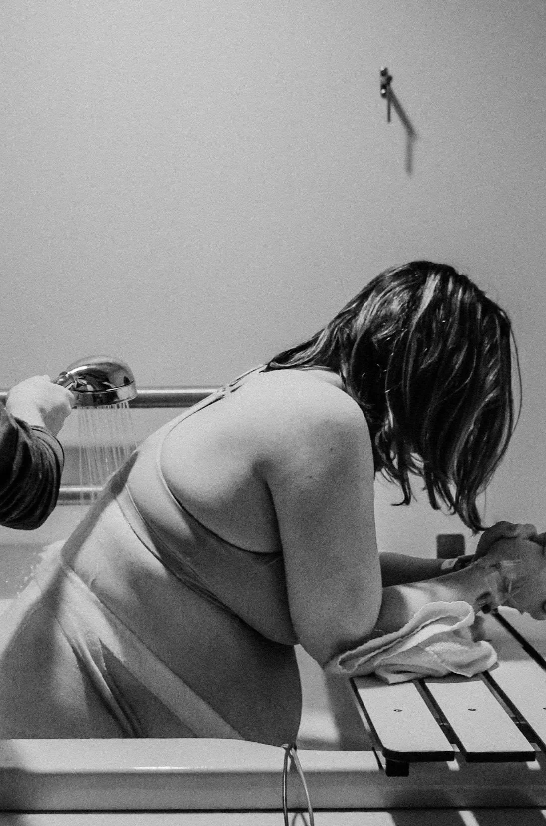 A woman in a swimsuit leaning over a poolside table, with a person holding a showerhead nearby. The scene appears to be in a locker room or pool area, in black and white.