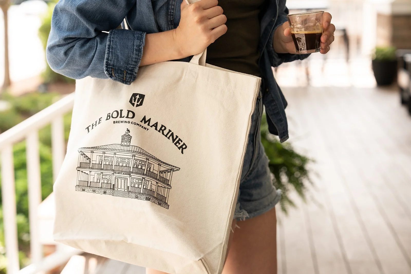 Woman holding an espresso shot and The Bold Mariner tote bag on porch