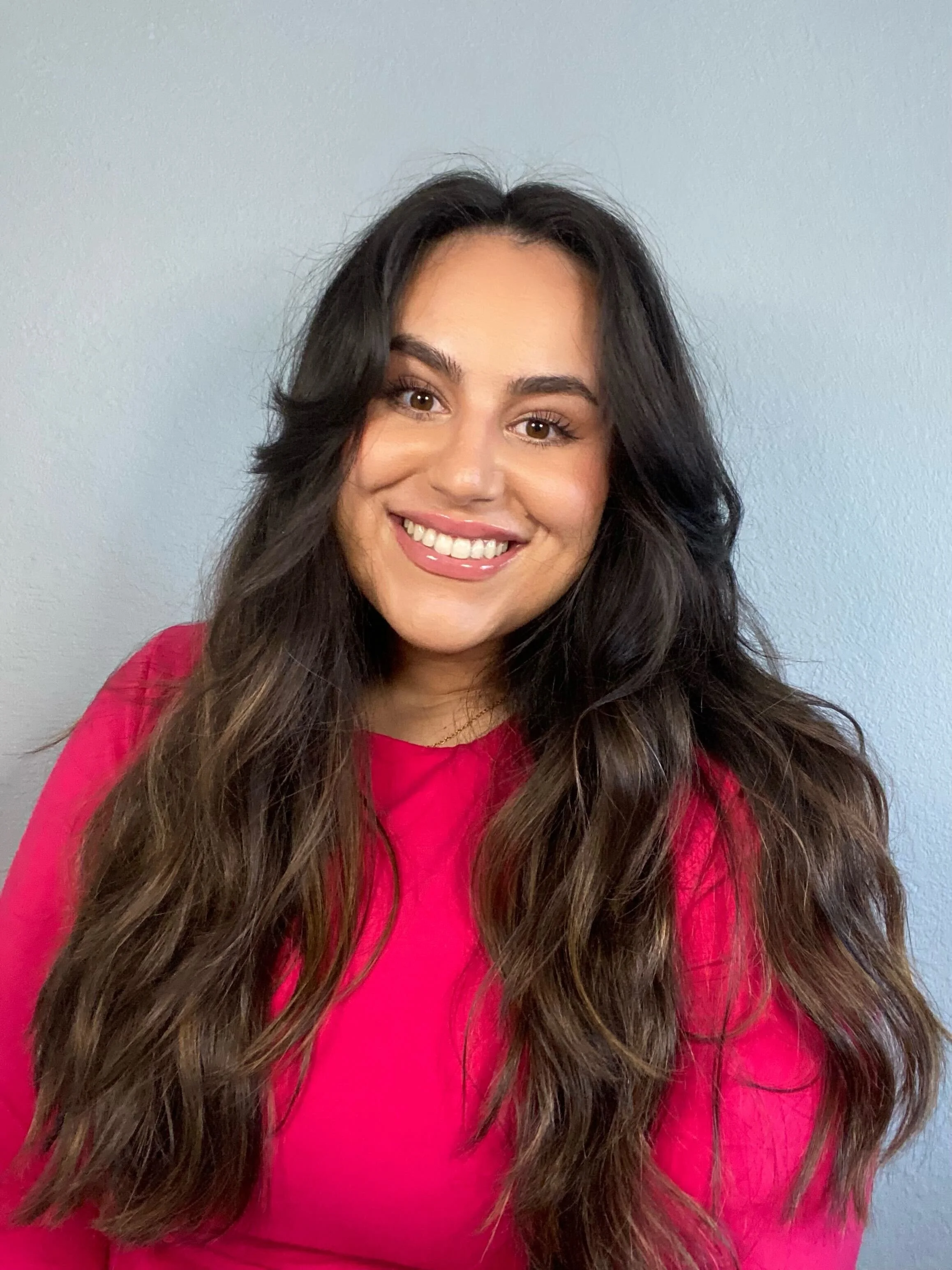 A smiling woman with long, wavy dark hair wearing a bright pink top, standing against a plain light gray wall.