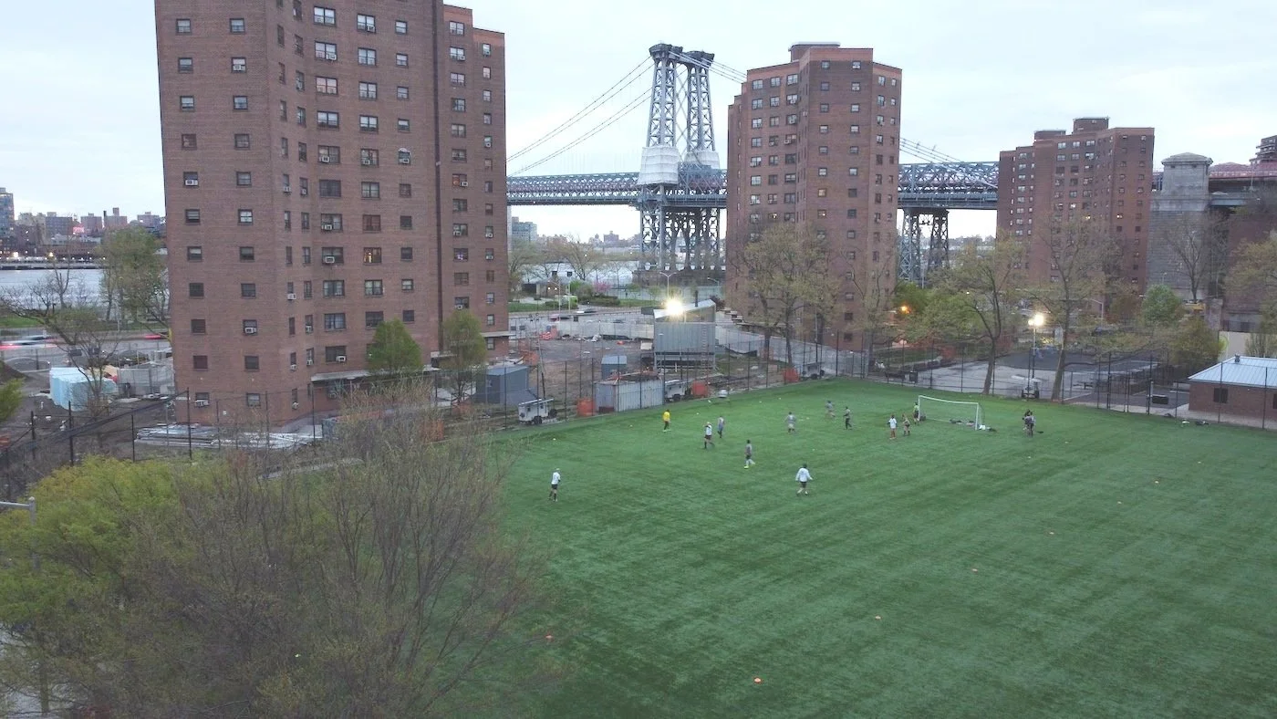 NYC Footy LES urban soccer field with players, surrounded by residential buildings and a bridge in the background.