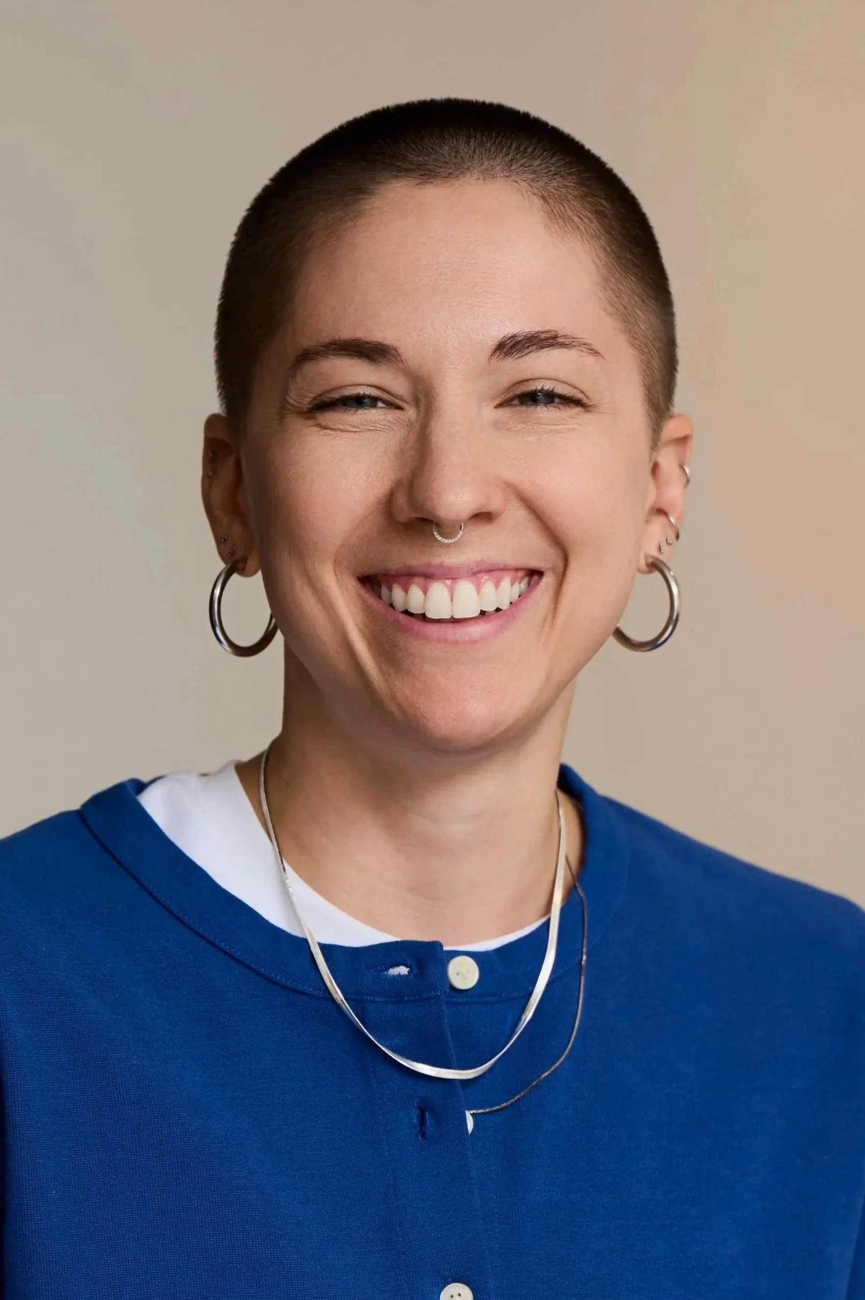 Portrait of a smiling person with a buzz cut, wearing large hoop earrings, a septum piercing, and layered necklaces, dressed in a blue top with a white undershirt, against a plain background.