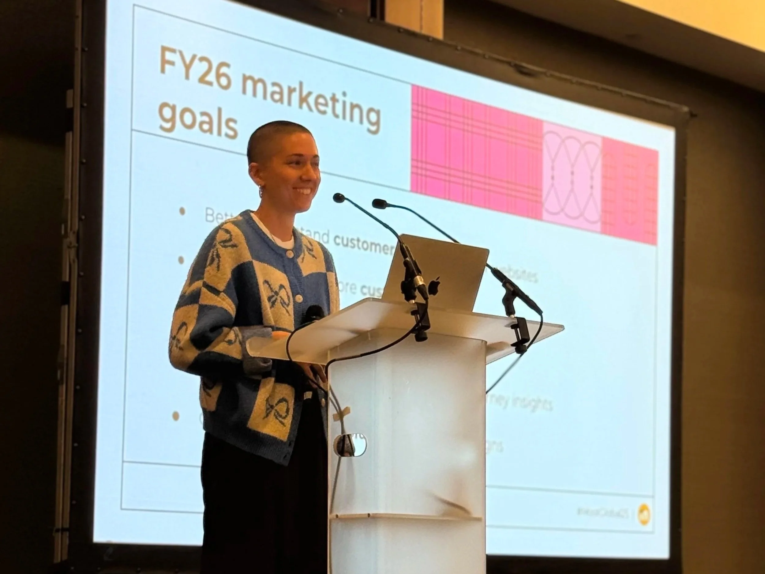 A person with a shaved head standing at a podium in front of a presentation slide titled 'FY26 marketing goals'. They are smiling and wearing a patterned sweater. There are two microphones on the podium and a laptop.