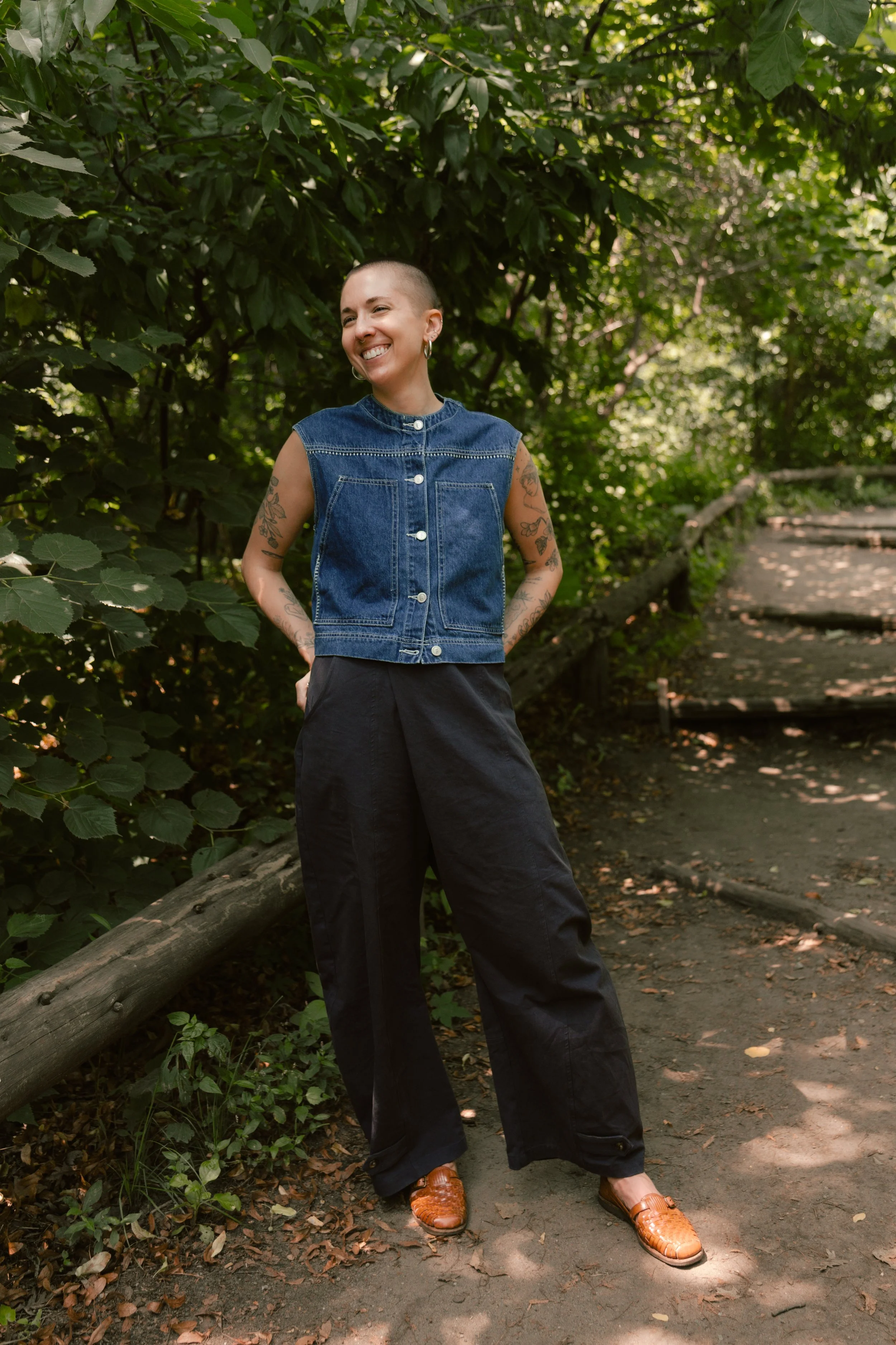 A photo of a person with a buzz cut, blue denim vest, blue trousers, in a park