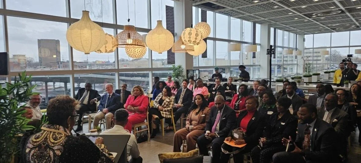 A diverse group of people seated in a modern conference room with large windows, listening to a speaker at a panel discussion or seminar. The room is decorated with hanging lamps and potted plants.