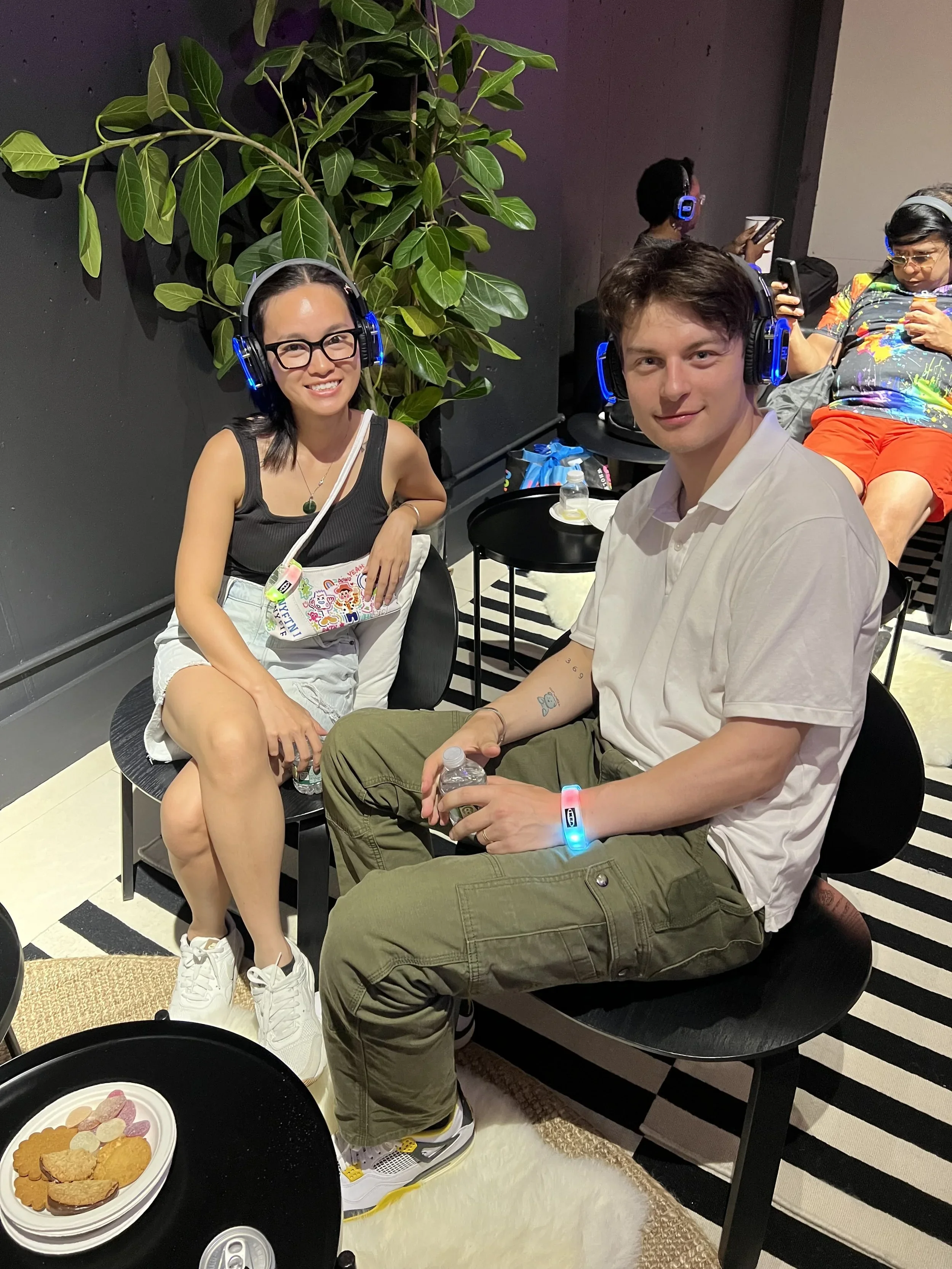 Two people sitting at a table with snacks, wearing headphones, smiling, and looking at the camera, with others in the background also wearing headphones and using their phones, a large green plant and a black wall behind them.