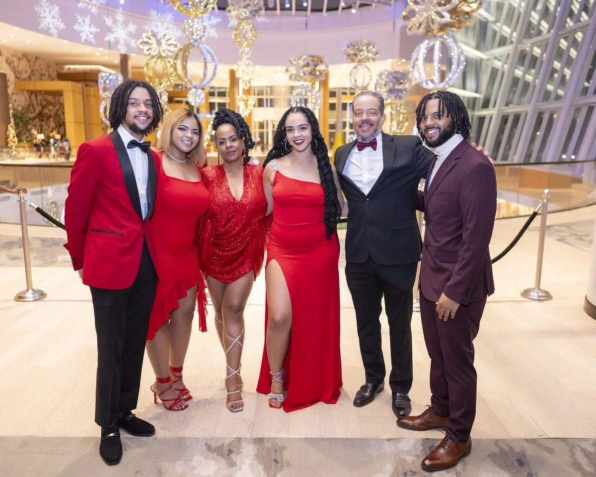 Six people dressed in formal attire standing together in a decorated, festive indoor setting with holiday ornaments and lights.