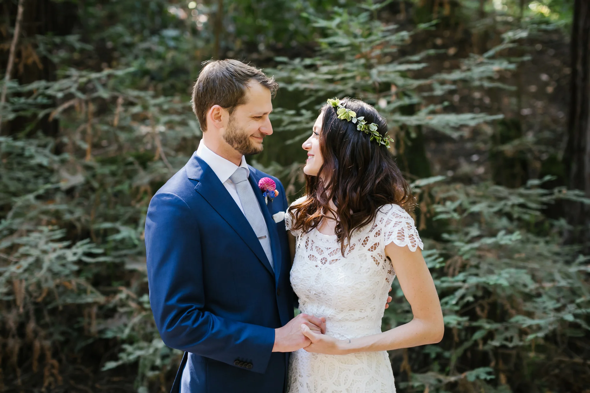Groom in blue suit holds hands with his bride in a lace dress and flower crown