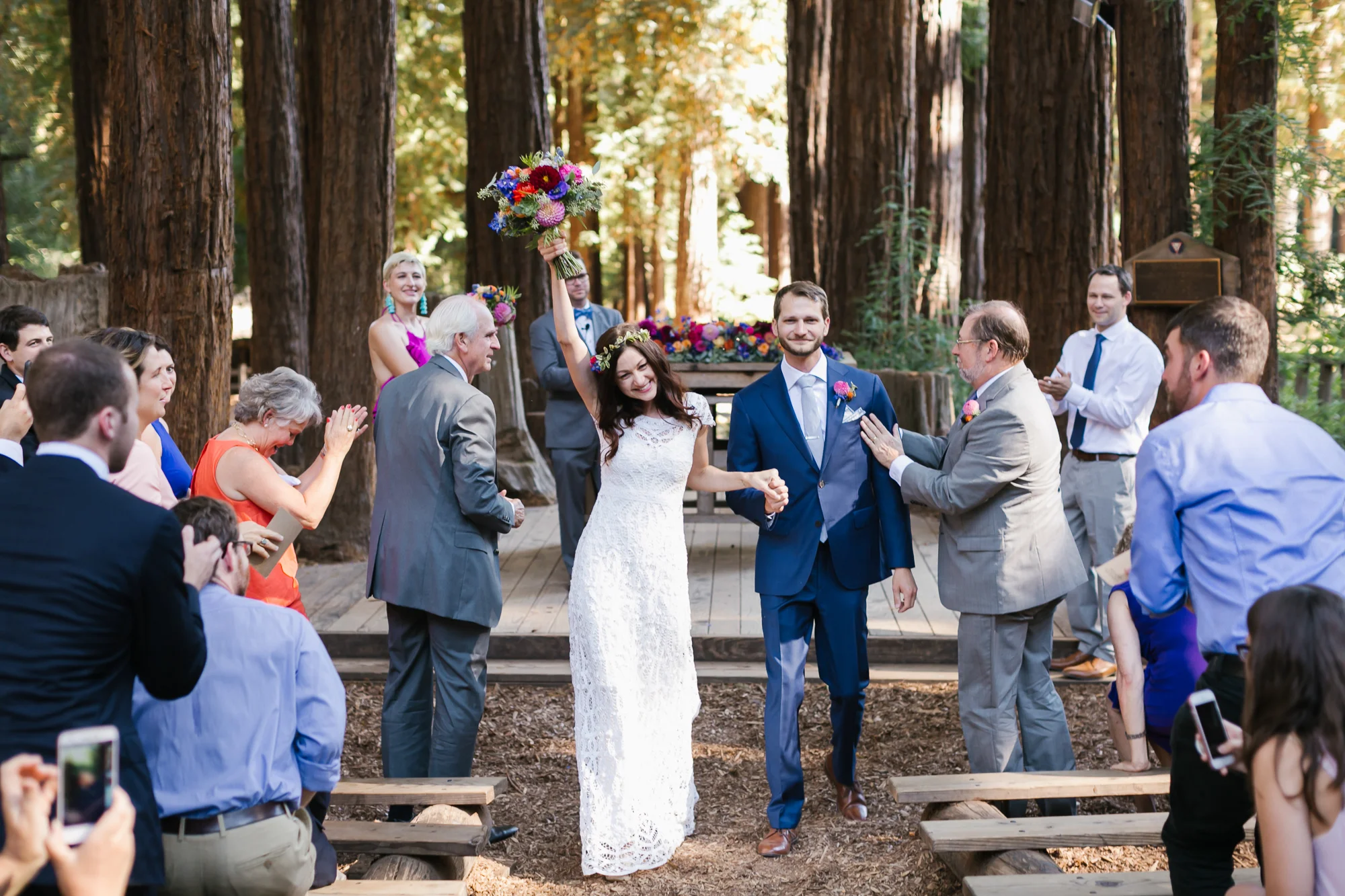 Bride holds her colorful bouquet up over her head in celebration as she walks out of ceremony with her husband