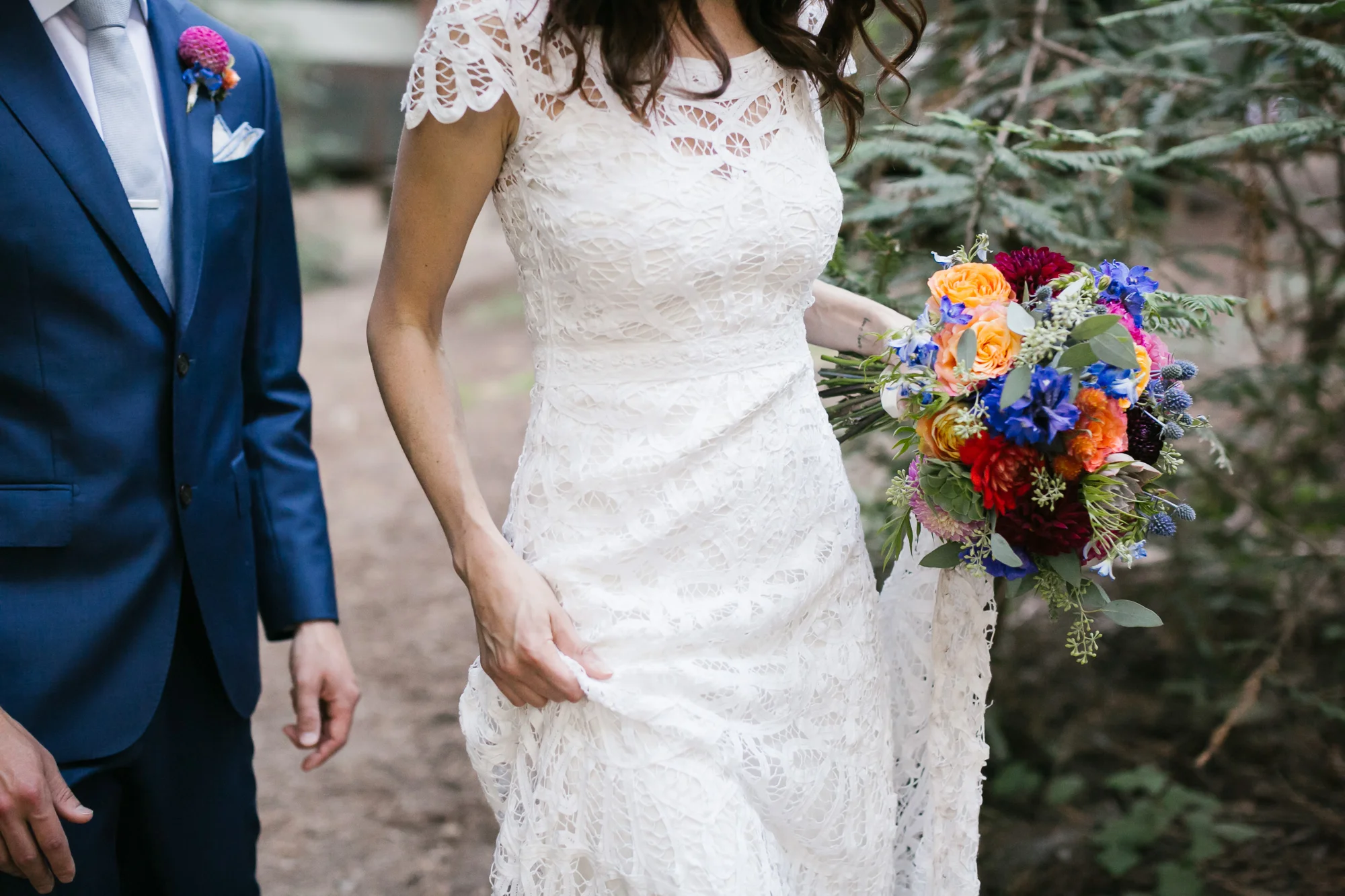 Bride holds her lace gown and bright bouquet as she walks with her husband in his blue suit