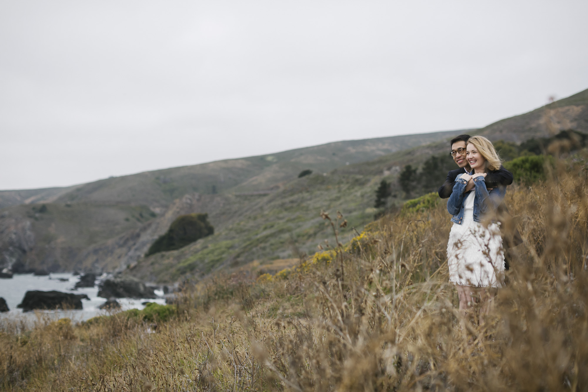 On a coastal hillside, a man hugs his fiancé from behind while looking out at the ocean