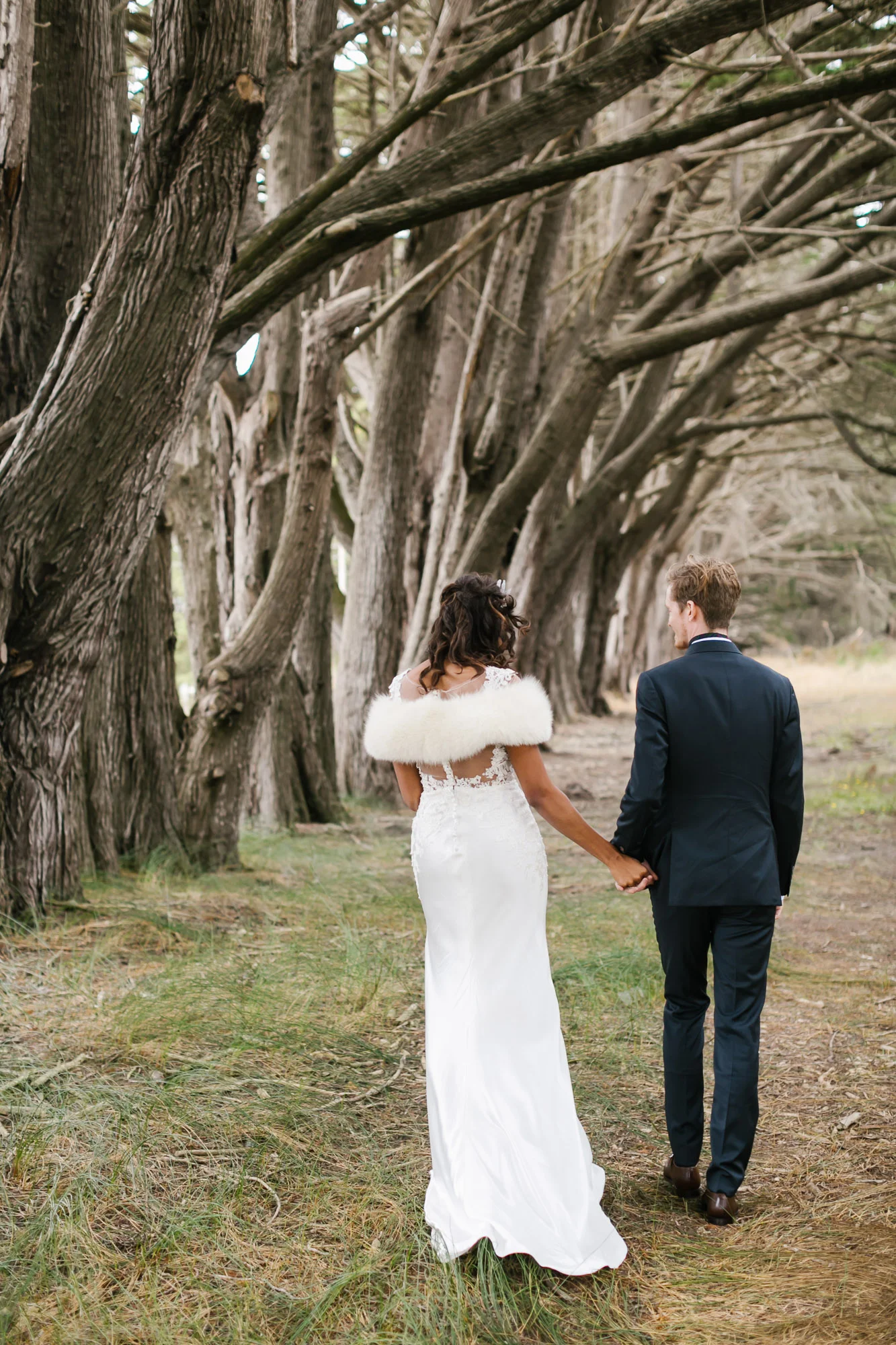 Bride wearing crystal crown walks with groom among coastal trees