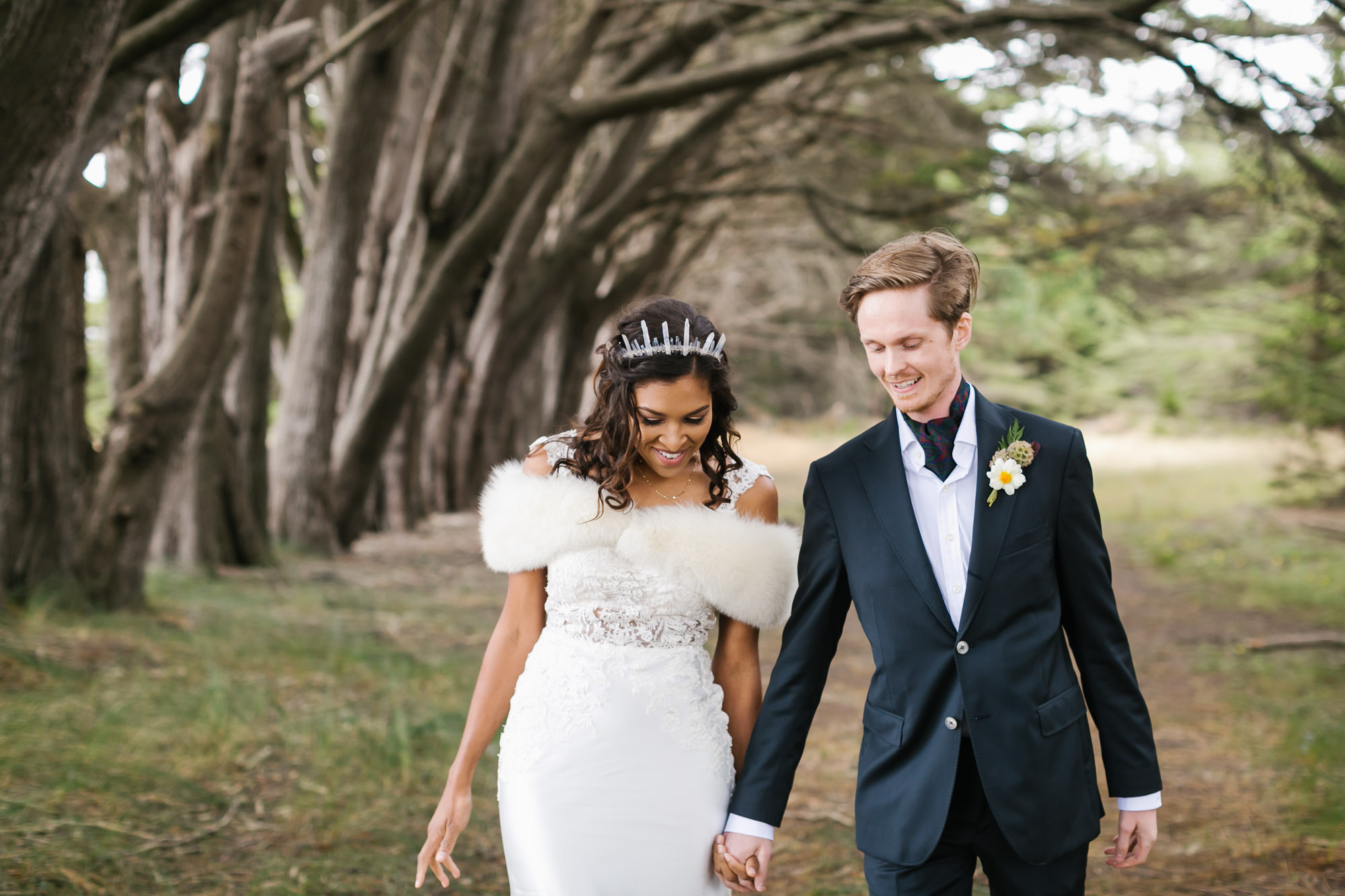 Bride wearing crystal crown and white fur stole walks holding hands with her husband