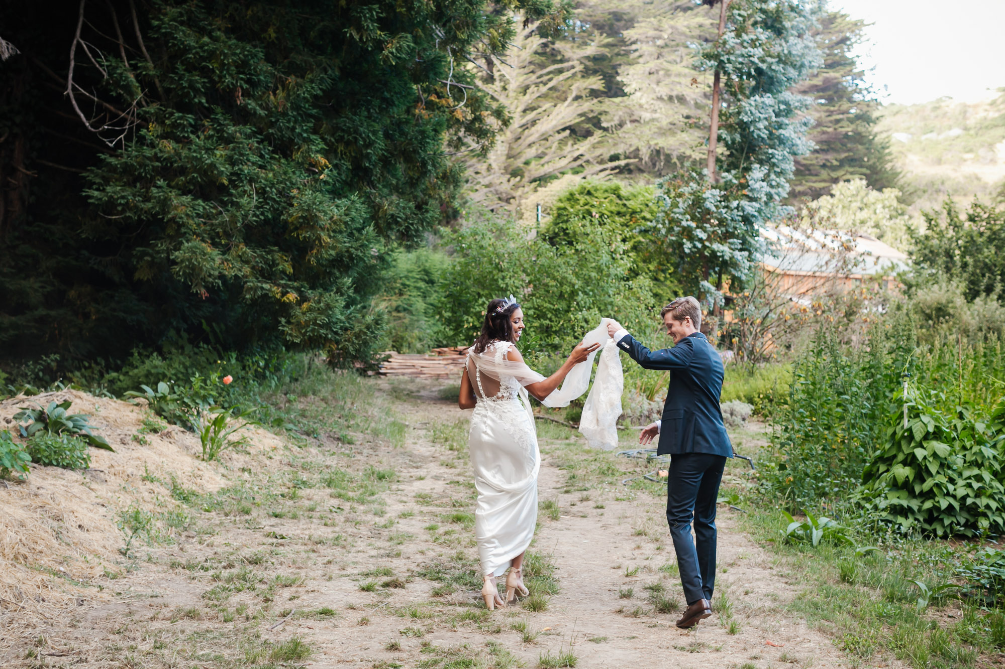 Groom helps his bride with her veil as they walk through coastal farm